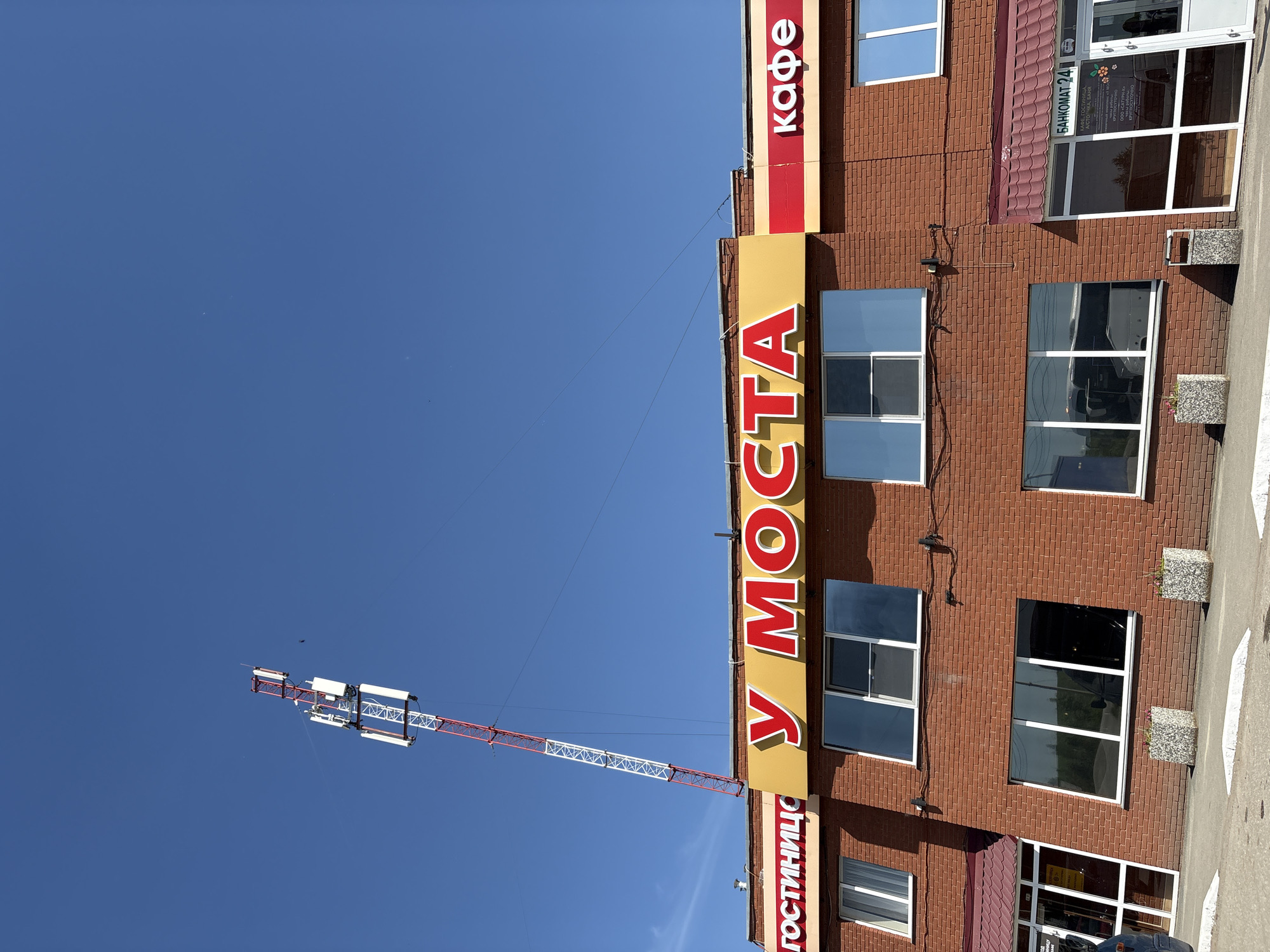 The image depicts a section of a brick building located near a bridge, possibly in the city of Grishanki, Russia. Vertical signs reading "ГОСТИНИЦЕ У МОСТА" (Hotel at the Bridge) and "кафе" (cafe) are affixed to the building's facade. A sign indicating "БАНКОМАТ 24" (ATM 24) is present, suggesting round-the-clock availability of banking services. The windows reflect the sky and surrounding environment. A construction crane is visible extending into the clear blue sky. The ground near the building appears to be a paved area.