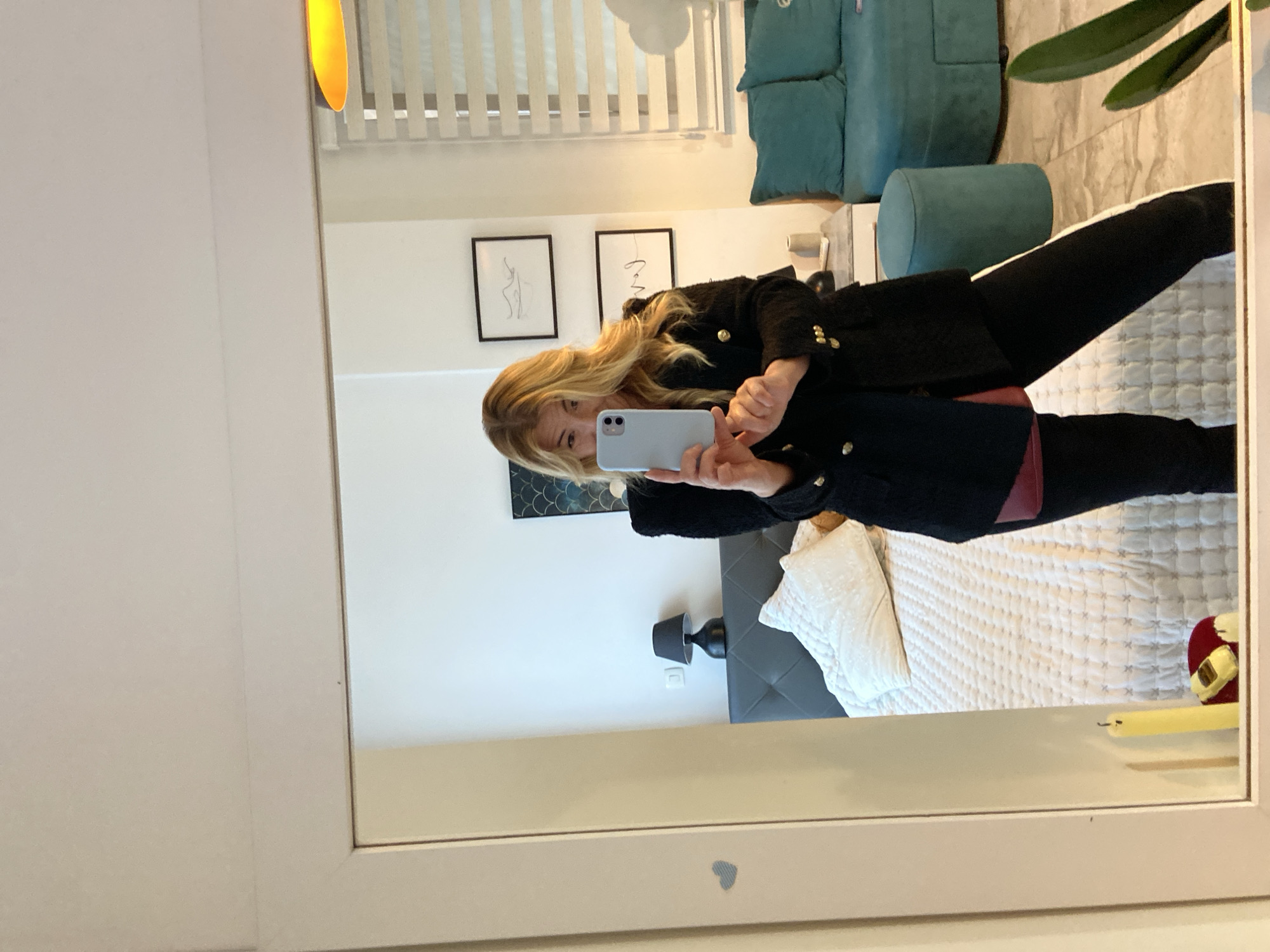 The image depicts a reflection in a mirror. A woman with blonde hair is holding a smartphone and appears to be taking a photo of herself in the mirror. She is wearing a black jacket with gold buttons and black leggings. The background reveals a bedroom setting. The scene includes a bed with white linens and a grey headboard, a turquoise couch and ottoman, artwork on the walls, a table lamp, and blinds on a window. A houseplant sits near the mirror. While the location is not explicitly discernible from the visual data provided, the city mentioned is Budva, Montenegro.