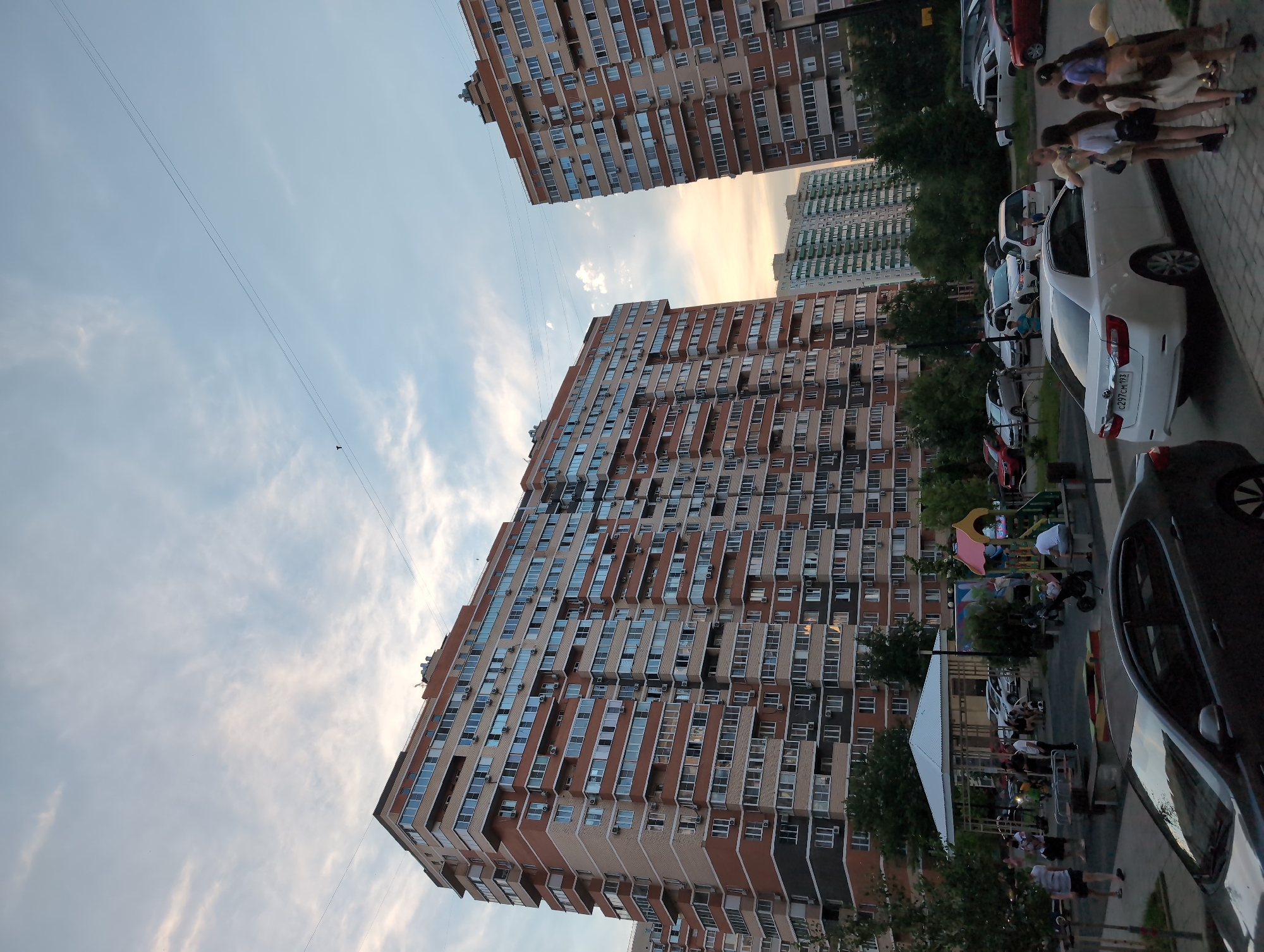 This image captures a street scene in Krasnodar, Russia, likely during the late afternoon or early evening. The setting is residential, dominated by tall, multi-story apartment buildings with numerous windows and balconies. In the foreground, a street is lined with parked cars, including a white sedan and a dark-colored vehicle. A group of people, including adults and children, are visible near the sidewalk and a playground area. Some individuals are walking, while others appear to be socializing. The playground features colorful structures, suggesting it is a family-friendly neighborhood. The sky is partly cloudy with a warm, soft light, indicative of sunset. The overall atmosphere appears relaxed and communal, with residents enjoying the outdoor space. A license plate on the white car reads "C297CM 193".