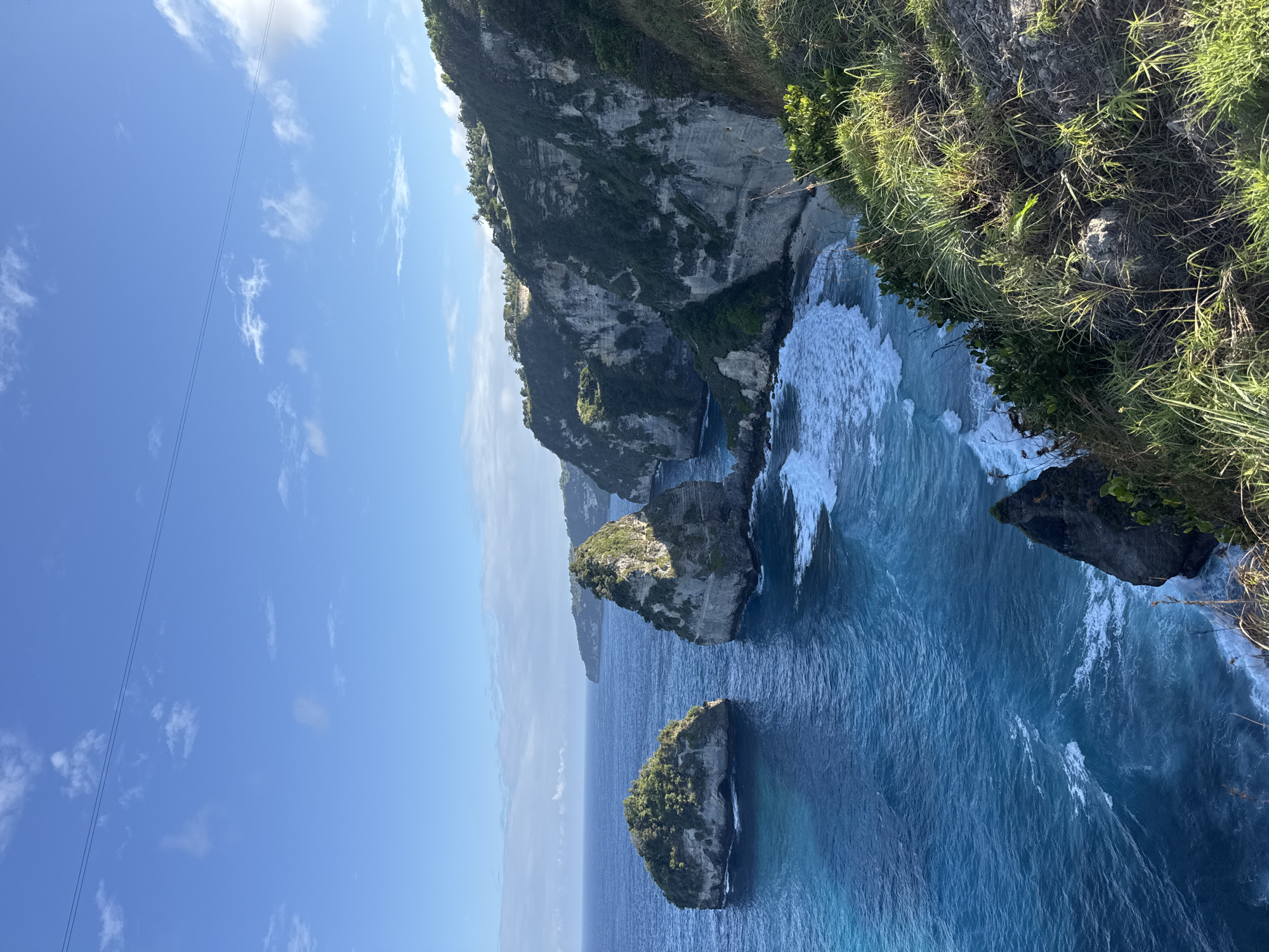 This is a medium shot, taken from a high vantage point, of a rocky coastline with turquoise water. The scene is dominated by the ocean and dramatic cliffs, with lush green vegetation clinging to the slopes. Several large rock formations rise from the sea, topped with verdant growth. Waves crash against the shore and the base of the cliffs, creating white foam. The sky is a clear blue with a few wispy clouds. In the distance, more coastline is visible, suggesting a vast, natural landscape. The image appears to be a scenic view of Nusa Penida, Indonesia, likely a popular tourist spot due to its breathtaking natural beauty. There are no people or man-made structures clearly visible in the immediate foreground or on the rock formations, although some structures might be present on the distant clifftops. The overall impression is one of rugged, natural splendor under a bright sky.