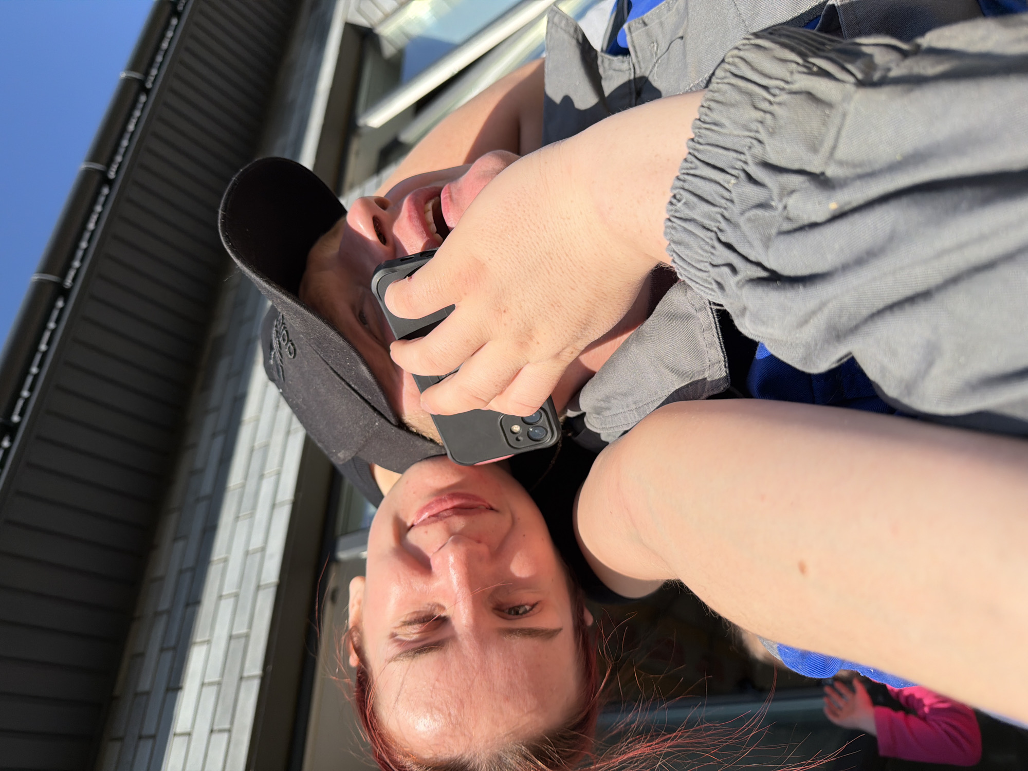 A person is holding a black mobile phone to their right ear while looking down at the camera. They are wearing a black cap and a grey jacket. The background shows a building exterior under a blue sky. The location is Chaikovsky, Russia.