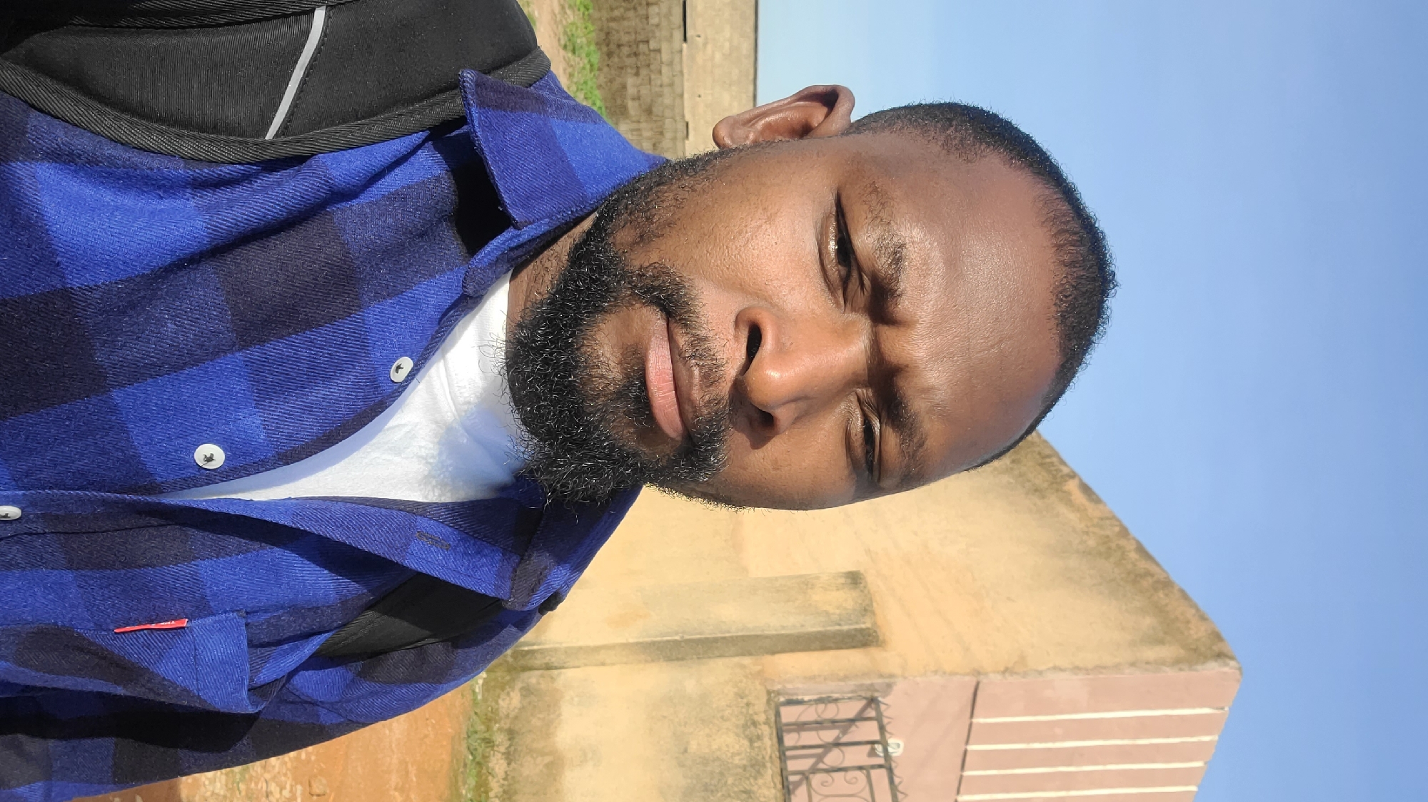 This is a portrait-style photograph of a Black man with a beard, wearing a blue and black checkered shirt over a white t-shirt. He has his eyes closed and his head is tilted back slightly, suggesting a relaxed or contemplative mood. The man is wearing a black backpack.
The background shows a building with light-colored walls and a metal grate on a window. The sky is clear and bright blue, indicating a sunny day. The lighting suggests it is daytime, likely morning or afternoon.
The image appears to be a selfie taken outdoors in a bright environment. The setting seems to be an outdoor area near a building, possibly in a residential or public space. There is no visible text in the image.