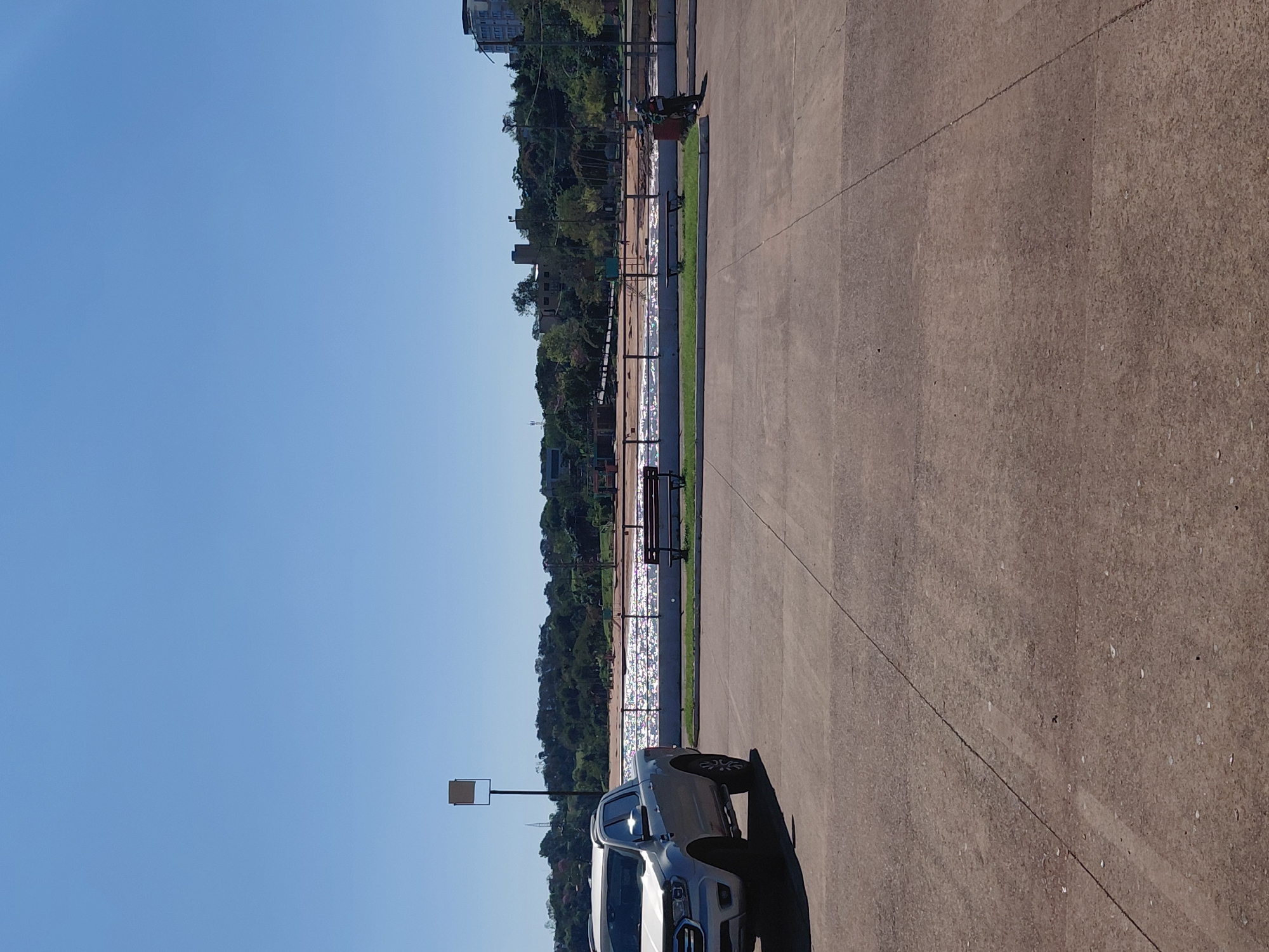 The image shows a view of the Costanera in Encarnación, Paraguay, on a clear, sunny day. In the foreground, a silver pickup truck is parked on a paved area. Beyond the paved area, there is a grassy strip with a wooden bench. Further back, a body of water, likely the Paraná River, sparkles under the sun, with a promenade or walkway visible along its edge. The opposite bank of the river is lined with lush green trees and some buildings, including a tall, modern structure. A person on a motorcycle is visible near a trash receptacle on the right side of the frame. The sky is a clear, bright blue.