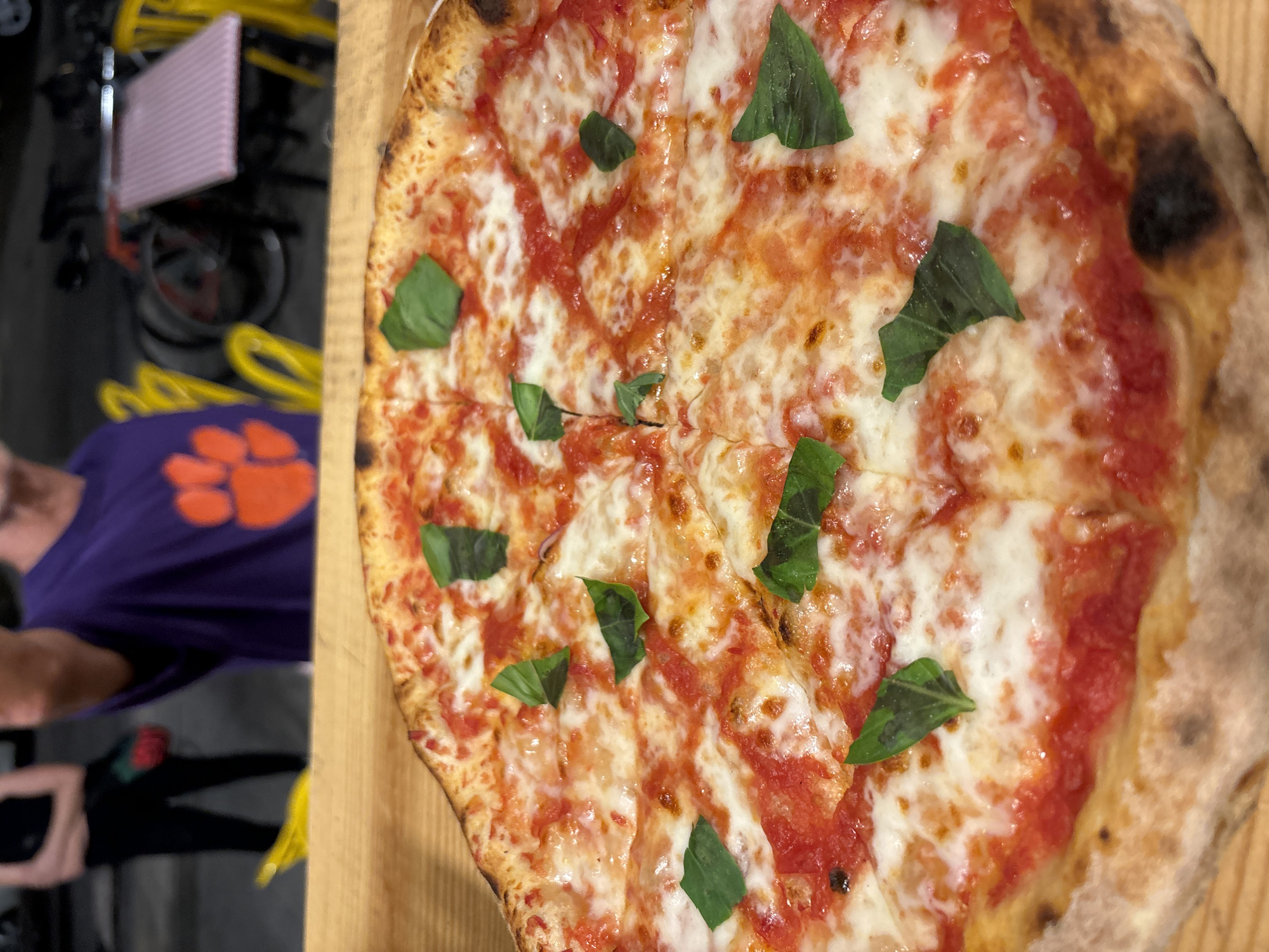The dominant subject of the image is a freshly baked Neapolitan-style pizza, centrally placed on a light-colored wooden board. The pizza is round, generously topped with bright red tomato sauce and melted, creamy white mozzarella cheese, characteristic of a Margherita pizza. Several vibrant green basil leaves are scattered across the surface. The crust exhibits classic "leopard spots" of charring, indicating a high-temperature bake. The pizza is already cut into multiple slices, suggesting it's ready to be served or eaten.
In the blurred background to the left, partially visible, is a person wearing a purple short-sleeved shirt with a prominent orange logo that resembles the paw print symbol of Clemson University. Below the person's upper body, other objects are indistinct but include coiled yellow cables and a pink, ribbed rectangular item. The overall setting appears to be indoors, possibly a casual dining area or kitchen. No specific time of day or weather conditions are discernible, and no emotions are clearly conveyed. Beyond the university logo, there is no visible text.