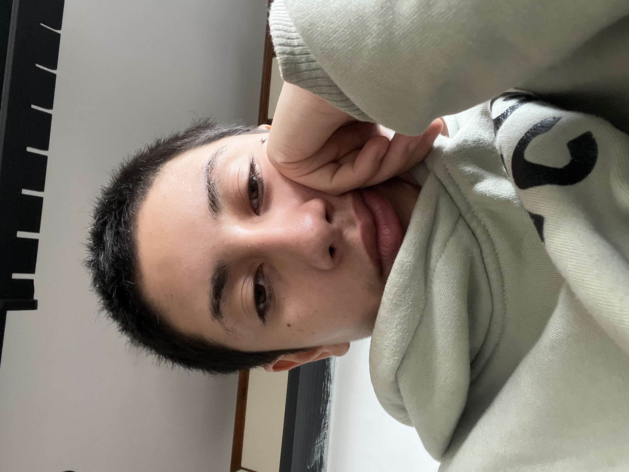 A close-up selfie of a young person with short dark hair and brown eyes, resting their head on their hand. They are wearing a light green hooded sweatshirt. The background is out of focus, but appears to be an indoor setting with a light-colored wall and a dark ceiling fixture. There is a framed picture on the wall in the background. There is no indication of the location in the city of Buenos Aires, Argentina.
