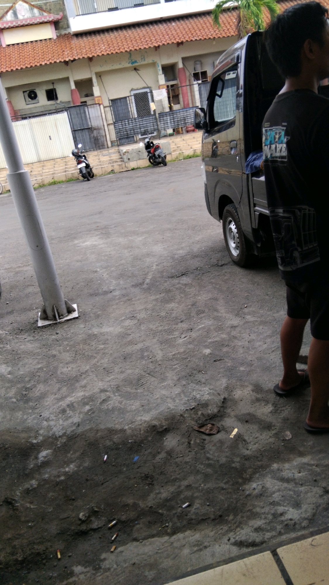 The image shows a street scene in Pekalongan, Indonesia. In the foreground, a person wearing shorts and flip-flops stands near a dark-colored pickup truck. The truck has a partial view of its side and rear. Behind the truck is a building with a red tiled roof, featuring windows and an air conditioning unit. Two motorcycles are parked on a raised walkway in front of the building. The ground is paved with asphalt and concrete, and there are several cigarette butts scattered on the ground in the lower part of the image. A metal pole, possibly a signpost or lamppost, stands in the left mid-ground. The lighting suggests it is daytime.