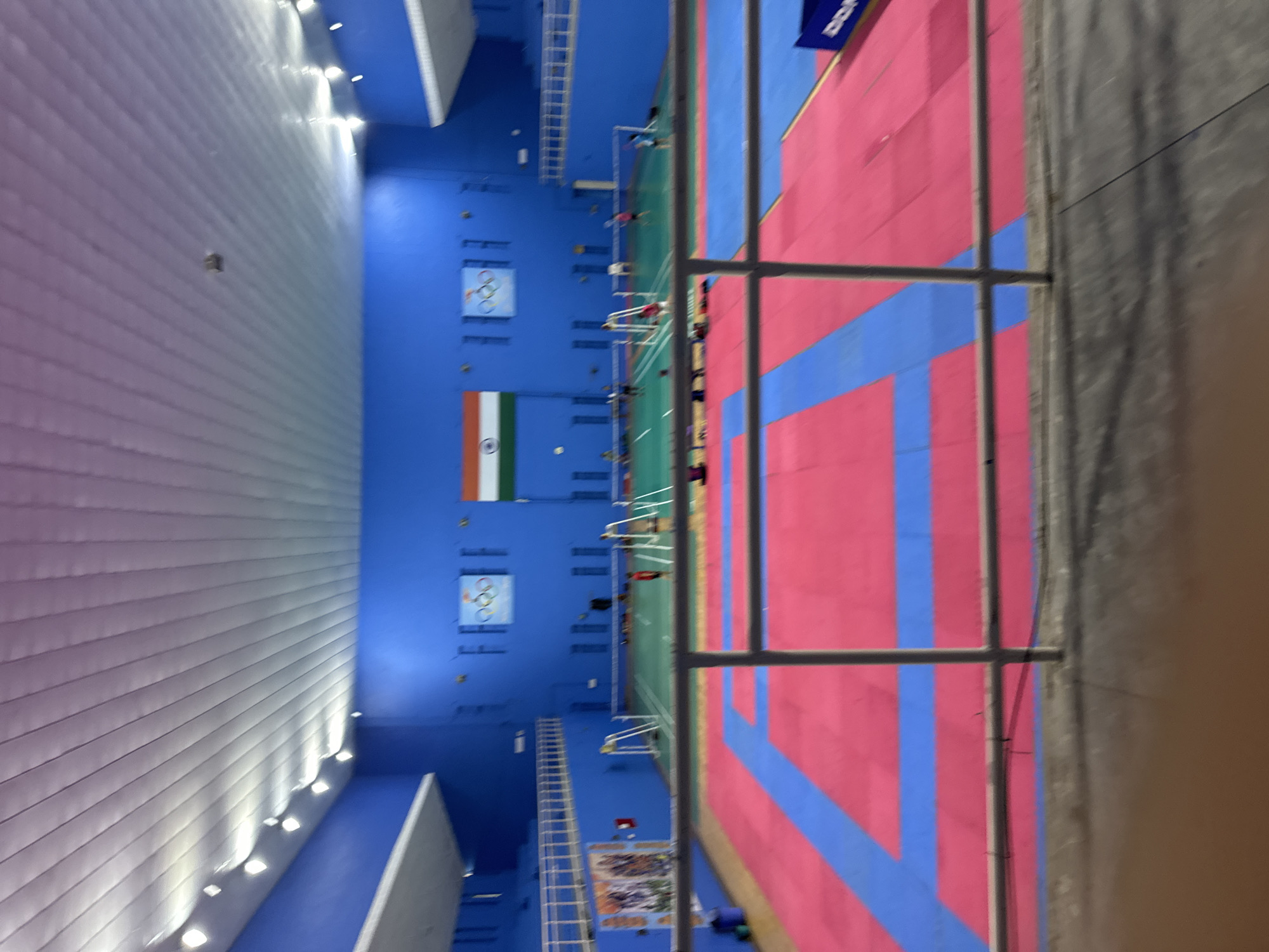 The image shows an indoor sports facility. The walls are painted a vibrant blue, and the ceiling is white with a ribbed texture and fluorescent lighting. In the background, the Indian flag is displayed on the wall, alongside Olympic rings. The floor of the facility is divided into several sections, with a large area of pink and blue mats, likely for martial arts or gymnastics. Further back, there are green badminton courts with nets and players visible. A railing is in the foreground, suggesting the photo was taken from a viewing area or elevated platform. The overall impression is of a well-maintained sports complex.