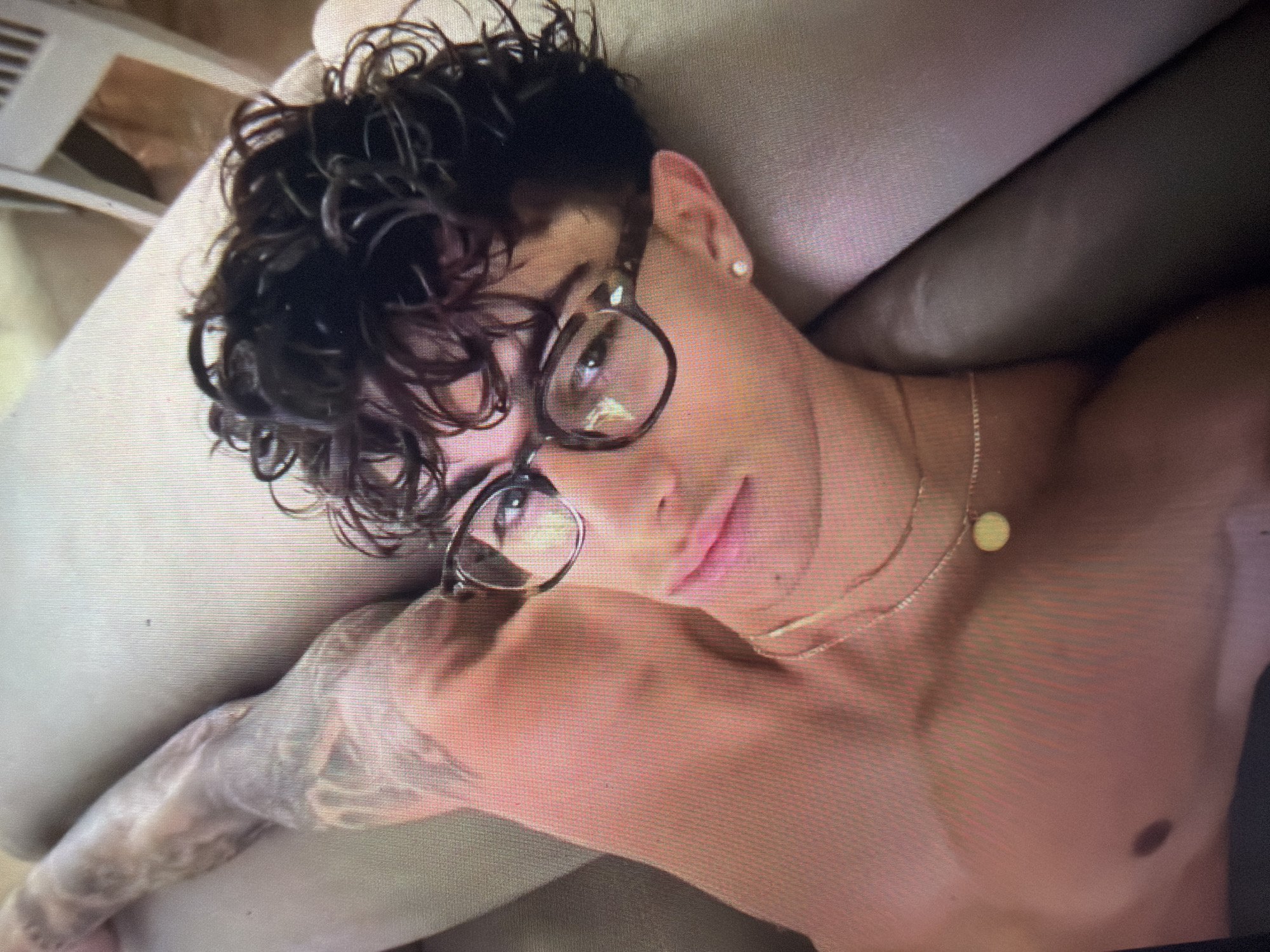 A youthful individual, appearing male, is depicted from the upper torso up, lying on their back on a light-colored soft surface, possibly a sofa or bed. The individual has dark, curly hair, dark-rimmed glasses, and a small, light-colored stud earring in the visible left ear. A gold-colored chain necklace with a small, round, flat pendant rests on the bare chest. Extensive tattooing is visible on the individual's left arm, appearing to be a full or partial sleeve. The individual's lips are slightly parted, and they are looking directly towards the viewer. The background is blurred, showing additional light-colored fabric and a portion of what appears to be a white slatted chair. The image exhibits a pixelated, screen-door effect, suggesting it may be a photograph of a digital screen. The scene is an interior setting. There is no visual information within the image to determine a specific geographic location such as the city of Shumen, Bulgaria.