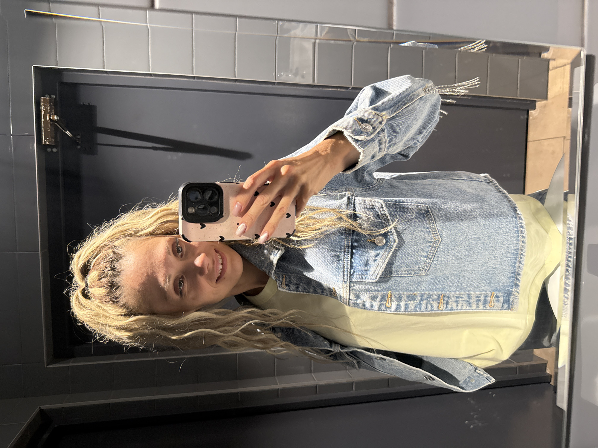 A young Caucasian woman with blonde, curly hair tied in a high ponytail is taking a mirror selfie in what appears to be a restroom. She is wearing a light yellow t-shirt under a light blue denim jacket adorned with silver fringe on the sleeves. The woman is smiling, showing some teeth, and her phone, with a pink case decorated with small black hearts, is held up to capture the reflection. The background shows a dark gray door and tiled walls, suggesting an indoor public space. The presence of a mirror indicates she is likely in a bathroom or dressing area. There are no visible indications placing this scene specifically in Ignatovo, Russia.