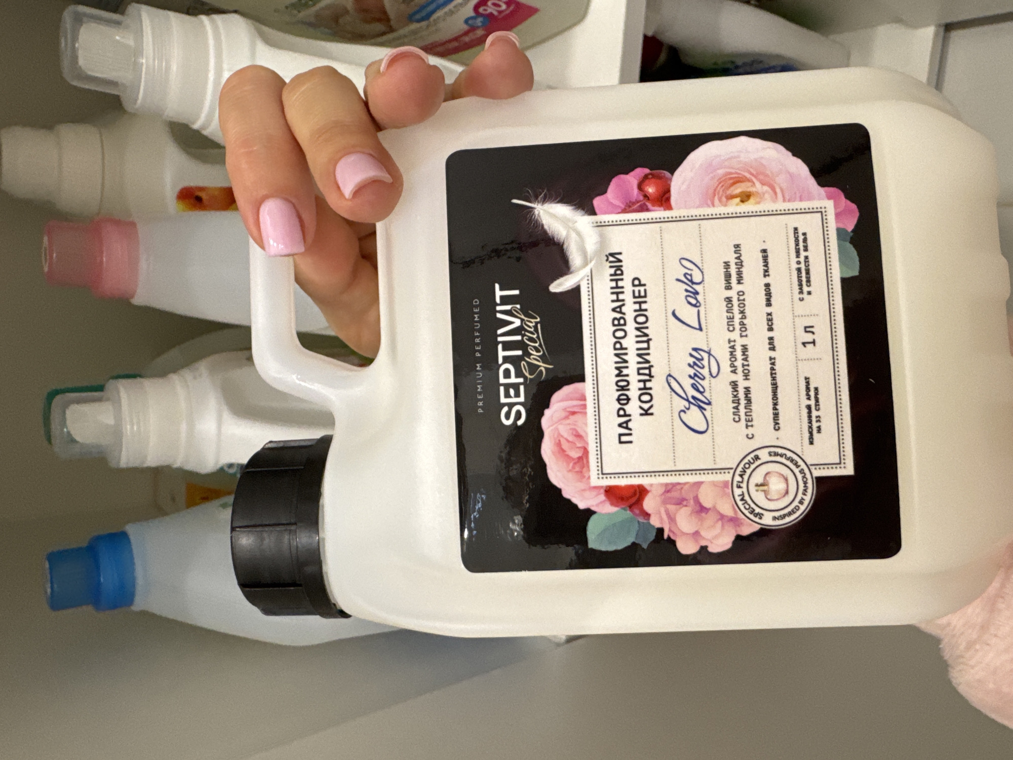 A person is holding a white plastic bottle of "SEPTIVIT Special" premium perfumed fabric softener, labeled "ПАРФЮМИРОВАННЫЙ КОНДИЦИОНЕР" and "Cherry Love." The bottle's label features a floral design with roses and mentions a "сладкий аромат спелой вишни с теплыми нотами горького миндаля" (sweet aroma of ripe cherry with warm notes of bitter almond). The bottle states it is a "суперконцентрат для всех видов тканей" (superconcentrate for all types of fabrics) and contains 1 liter.
The person's hand is visible, with manicured pink nails. In the background, other cleaning product bottles are visible. The image location is not explicitly identified as Sochi, Russia, although the product is likely from Russia based on the Cyrillic text on the label.