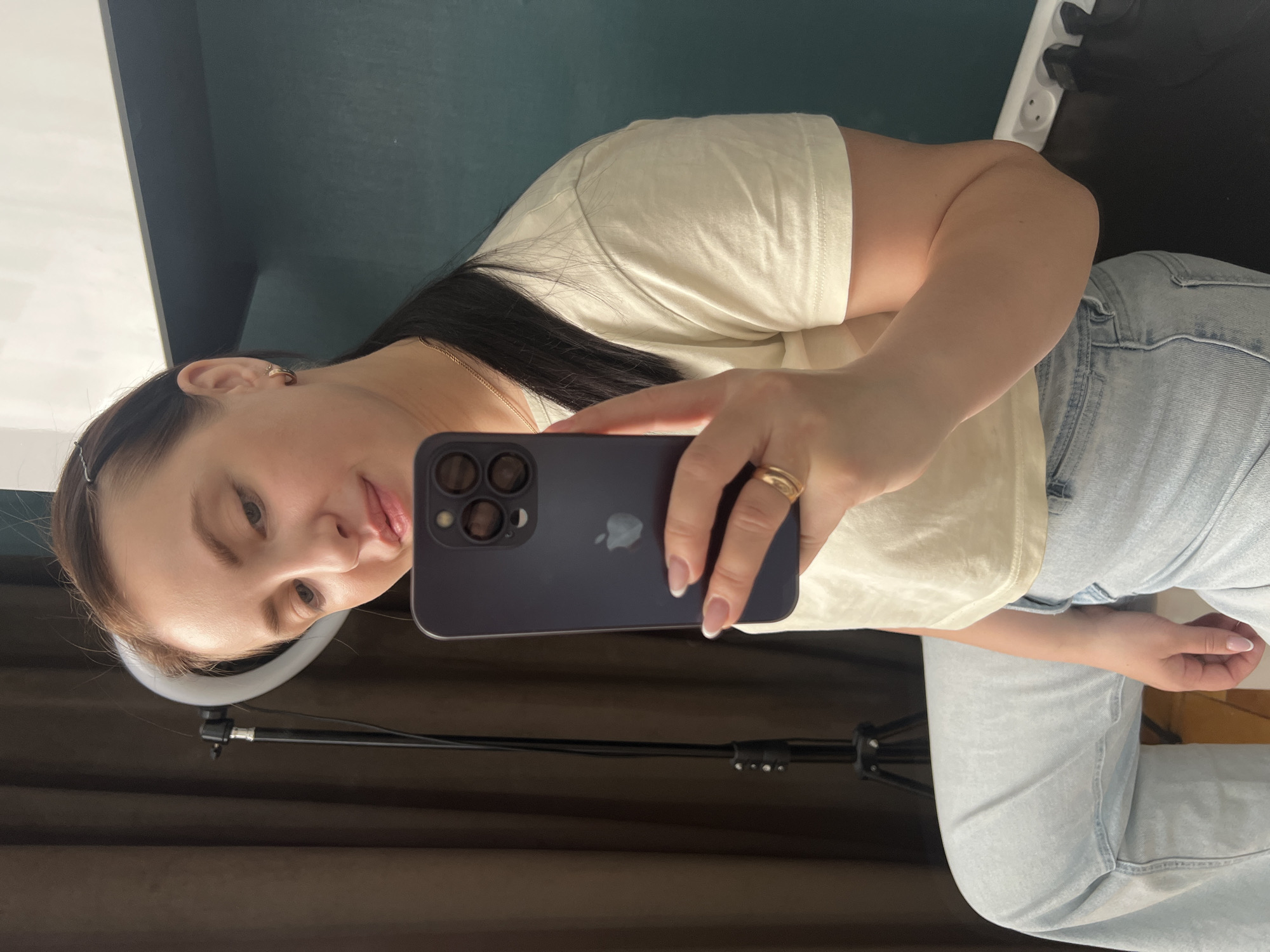 A woman is taking a selfie with a dark purple smartphone. She is wearing a beige t-shirt and light blue jeans. She has dark hair and a gold ring on her finger. A ring light is visible in the lower-left corner of the image. The background includes a dark curtain and what appears to be a wall with an electrical outlet. The location is Марковский, Russia.