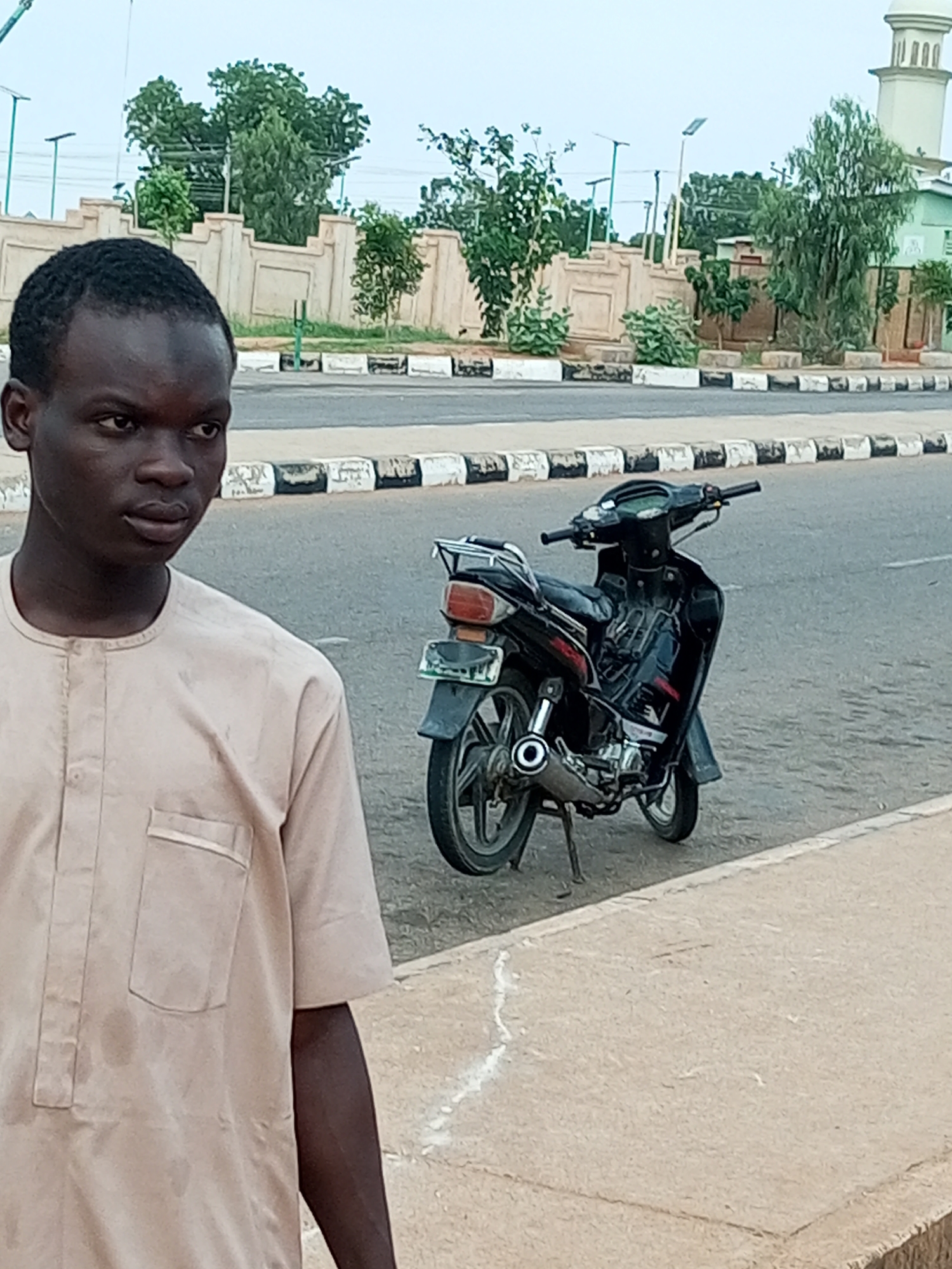 A young Black male, wearing a tan traditional garment with a pocket on the left chest, stands on a concrete sidewalk in Gombe, Nigeria. His dark hair is short and closely cropped. He is looking to his right, away from the camera. Behind him, a black motorcycle with a red taillight is parked on the asphalt road, positioned at an angle. The motorcycle has a kickstand engaged. The road is bordered by a sidewalk with a white line running along its edge. Further in the background, there is a decorative stone wall with arched elements and various trees. A white minaret of a mosque and a green building are visible in the distance. The sky is overcast.