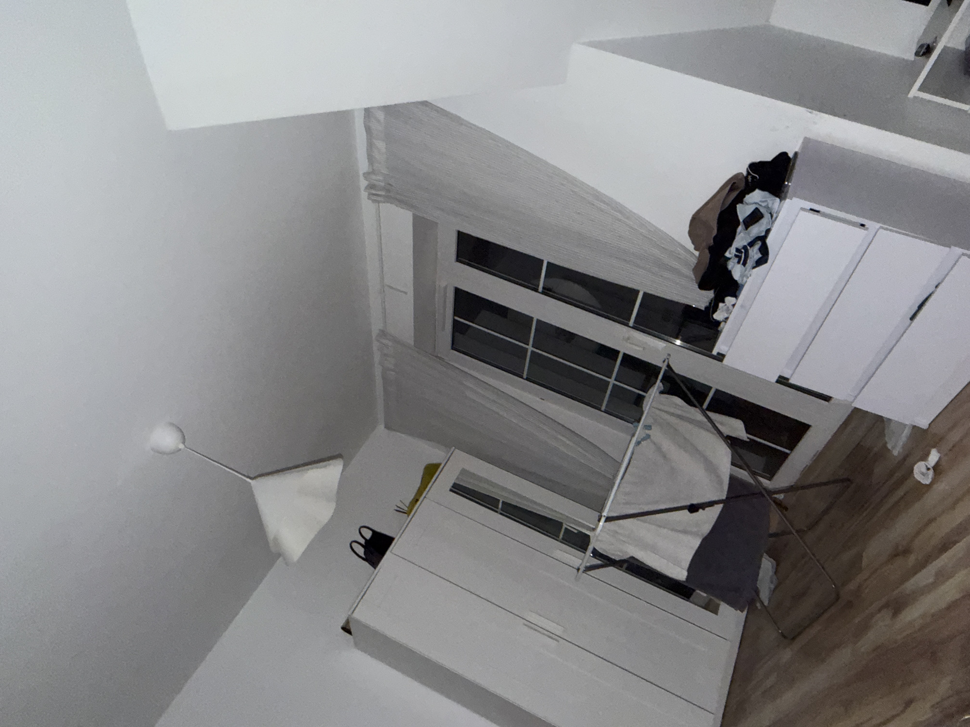 The image shows an indoor scene, likely a bedroom, in Falenty Nowe, Poland. The room is sparsely furnished and appears to be in a state of disarray. A large white wardrobe stands against the left wall. In the center of the room, a drying rack is set up with laundry draped over it. Behind the drying rack, a set of French doors with multiple panes leads outside. White sheer curtains partially obscure the doors. On the right side of the room, a chest of drawers is visible, with clothes piled on top. A ceiling light fixture hangs from the white ceiling. The floor is made of wood. The overall impression is one of a functional but untidy living space.