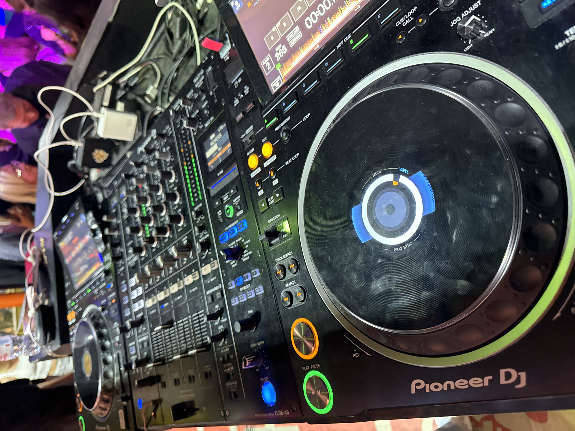 This image shows a Pioneer DJ DJM-A9 professional mixer. The mixer is black with a variety of knobs, buttons, sliders, and displays. The left side of the mixer contains multiple faders, knobs, and illuminated buttons for channel control. A digital display is visible above these controls. The center area features a crossfader and various control buttons, including track search and effect controls. On the right is a large jog wheel with a central screen showing "VINYL MODE". The mixer is illuminated with various colors, and there are connected cables and cords to the left of the device. The surrounding environment is dark, and blurred figures are visible. There's no information to indicate it's in Moscow, Russia.