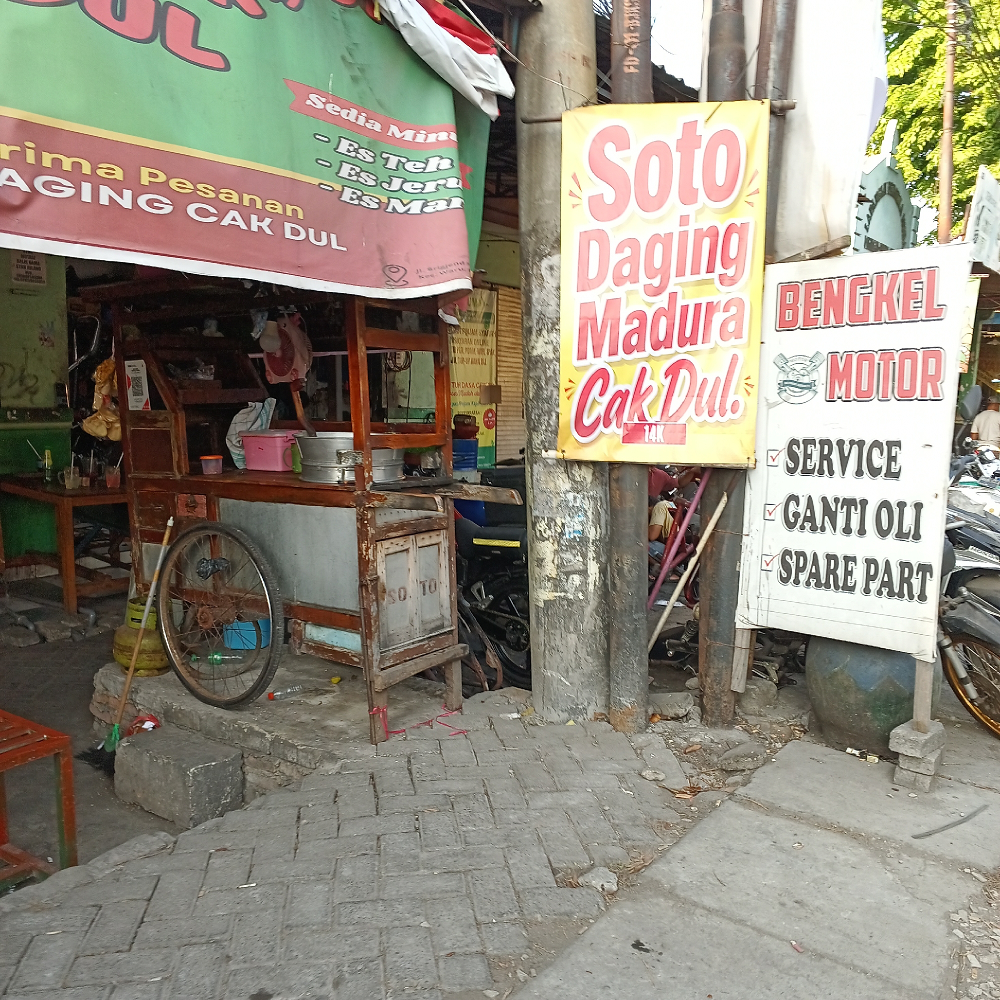 The image depicts a street scene in Wedoro, Indonesia, featuring a food stall and a motorcycle repair shop. In the foreground, a wooden food cart with various cooking utensils and containers is visible. A bicycle wheel and a gas cylinder are also present near the cart. Two prominent banners are displayed: one advertising "Soto Daging Madura Cak Dul" (Madurese Beef Soto by Cak Dul) and another for "BENGKEL MOTOR" (Motorcycle Workshop), listing services such as "SERVICE," "GANTI OLI" (Oil Change), and "SPARE PART." A section of the food stall's sign reads "terima Pesanan AGING CAK DUL" (accepting orders AGING CAK DUL). The background reveals a green wall with some graffiti and a glimpse of trees. The ground is paved with cobblestones, and the overall environment suggests a typical urban street setting in Indonesia. There are no visible people actively engaged in any activity, though the presence of the workshop implies ongoing business.