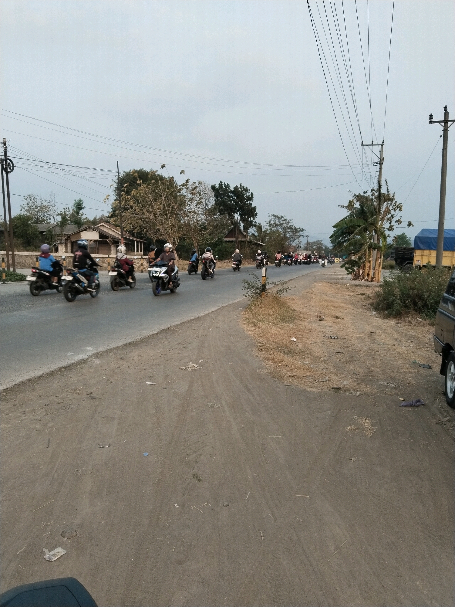 A convoy of motorcycles travels on a paved road in Songgom, Indonesia, under an overcast sky. The road is lined with dry grass and some scattered debris on the shoulder. Buildings with simple architecture and trees, some with sparse foliage, are visible behind the road. Utility poles with numerous wires are present along the roadside. A dark-colored vehicle, possibly a van, is partially visible on the right side of the image. Several people are riding motorcycles, most wearing helmets, suggesting travel or a group activity.