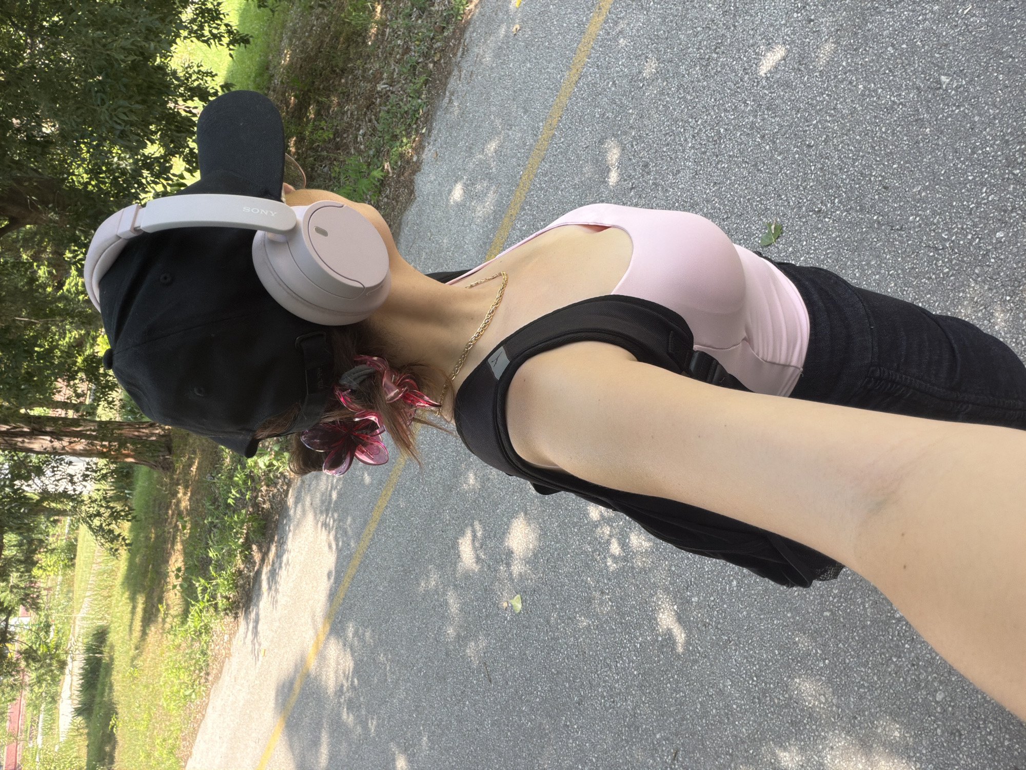 The image captures an individual from a high-angle, over-the-shoulder perspective, facing away from the camera while on a paved outdoor path. This scene is located in Pazardzhik, Bulgaria.
The individual wears a black baseball cap backward, under which dark hair is styled upwards, adorned with two transparent, magenta flower-shaped hair clips. Over the cap, light pink/beige over-ear headphones with "SONY" branding on the left earcup are worn. A gold-colored, chunky chain necklace is visible around the neck. The subject is dressed in a pink sleeveless top and dark-colored bottoms, with a black backpack worn on the shoulders, its straps clearly visible. The right arm is extended forward, consistent with holding a camera to capture the image, revealing fair skin with a small mole on the bicep.
The ground is a gray asphalt path with visible aggregate, marked by a single unbroken yellow line. To the left of the path, a grassy area with varied green foliage and smaller plants extends into the background, where a dirt track is also visible. To the right, dense tree cover with prominent tree trunks creates dappled shadows across the path. The scene is illuminated by bright natural sunlight, casting distinct shadows on the ground.