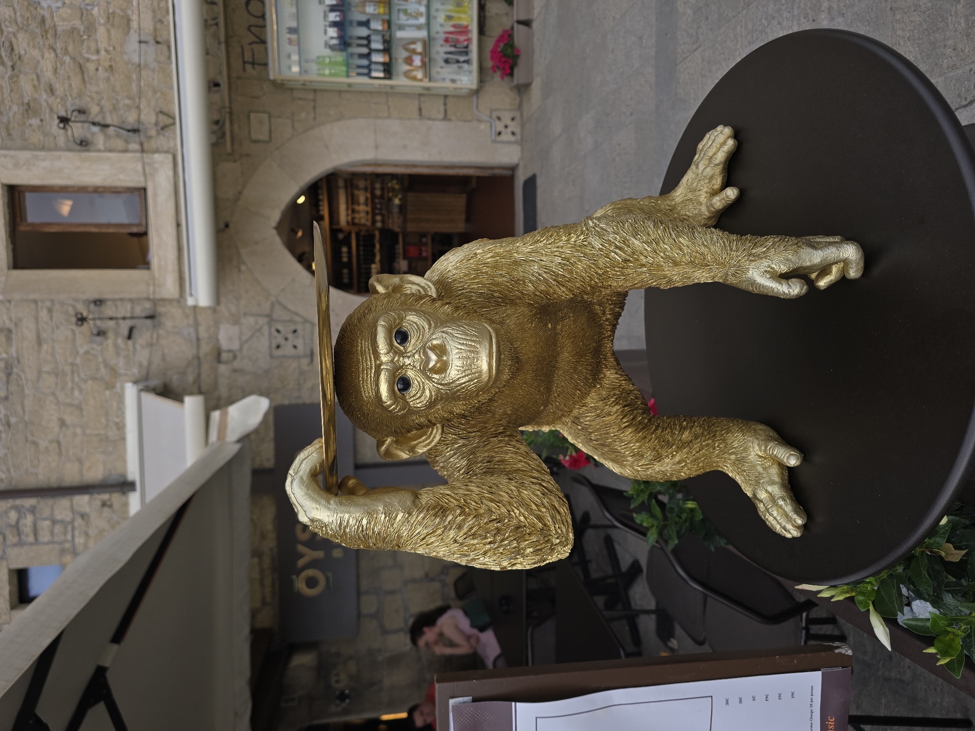 A golden monkey statue is posed in a crouching position on a dark round table, its left hand balanced on its head holding a flat, golden plate. The statue is positioned outdoors in front of a building with stone walls and an arched entryway. Behind the statue, an awning extends over a seating area with tables and chairs. To the right of the entryway, a display case filled with bottles is visible. The ground appears to be paved. The statue is the primary subject, with the background providing context of an outdoor dining or shopping area.