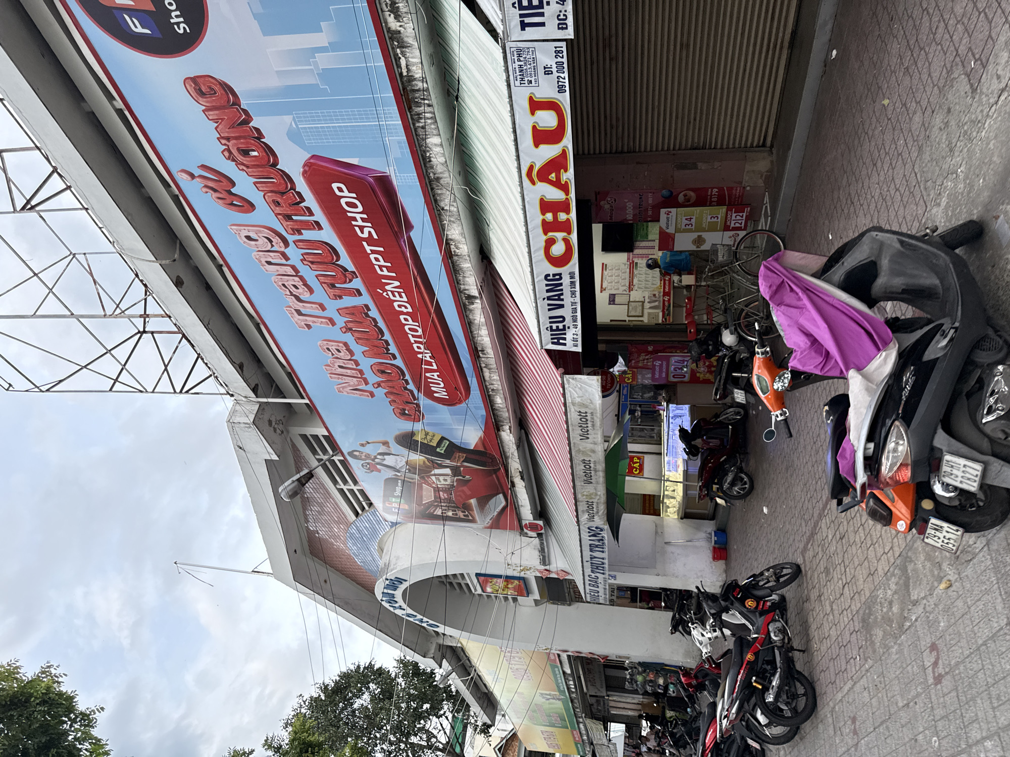 The image depicts a street scene in Nha Trang, Vietnam, likely in a commercial area. In the foreground, several motorcycles are parked on a brick-patterned sidewalk. A prominent billboard on the building facade displays advertising in Vietnamese for "FPT SHOP," featuring text like "NHÀ TRANG VI" and "CHÀO MÙA TỰU TRƯỜNG." The building has multiple storefronts, with awnings providing shade. One storefront is identified as "HIỆU VÀNG CHÂU," suggesting a jewelry store. Another sign indicates "TIỆC," potentially related to events or catering. A person in a blue shirt is visible inside one of the establishments. The overall environment appears to be a moderately busy urban setting on an overcast day.
