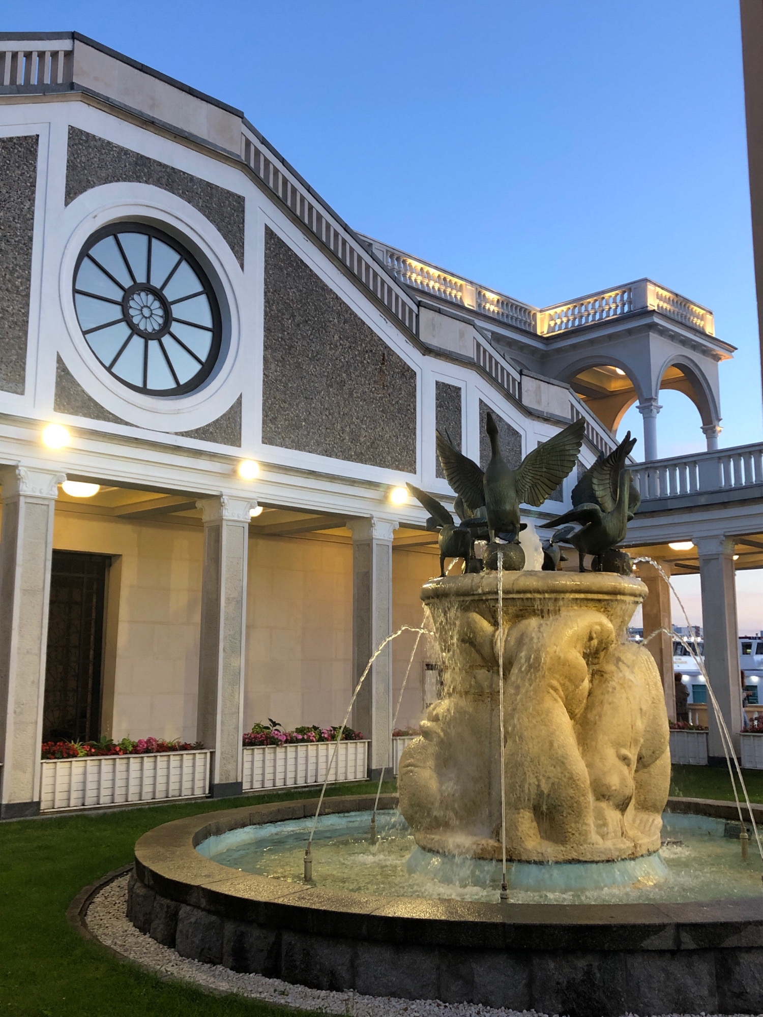 The image depicts the facade of a building with a fountain in the foreground. The building has a multi-level design with arched walkways and a decorative balustrade illuminated by lights. A prominent circular window with a radial design is visible on the upper part of the facade. The fountain features a sculptural element with several bird-like figures, possibly geese or swans, and a stone base from which water sprays. The surrounding area includes a well-maintained lawn, flower beds in white planters, and a paved walkway. The sky is clear and blue, suggesting it is dusk or dawn. In the background, a boat or ship is partially visible. This scene is located in Khimki, Russia.