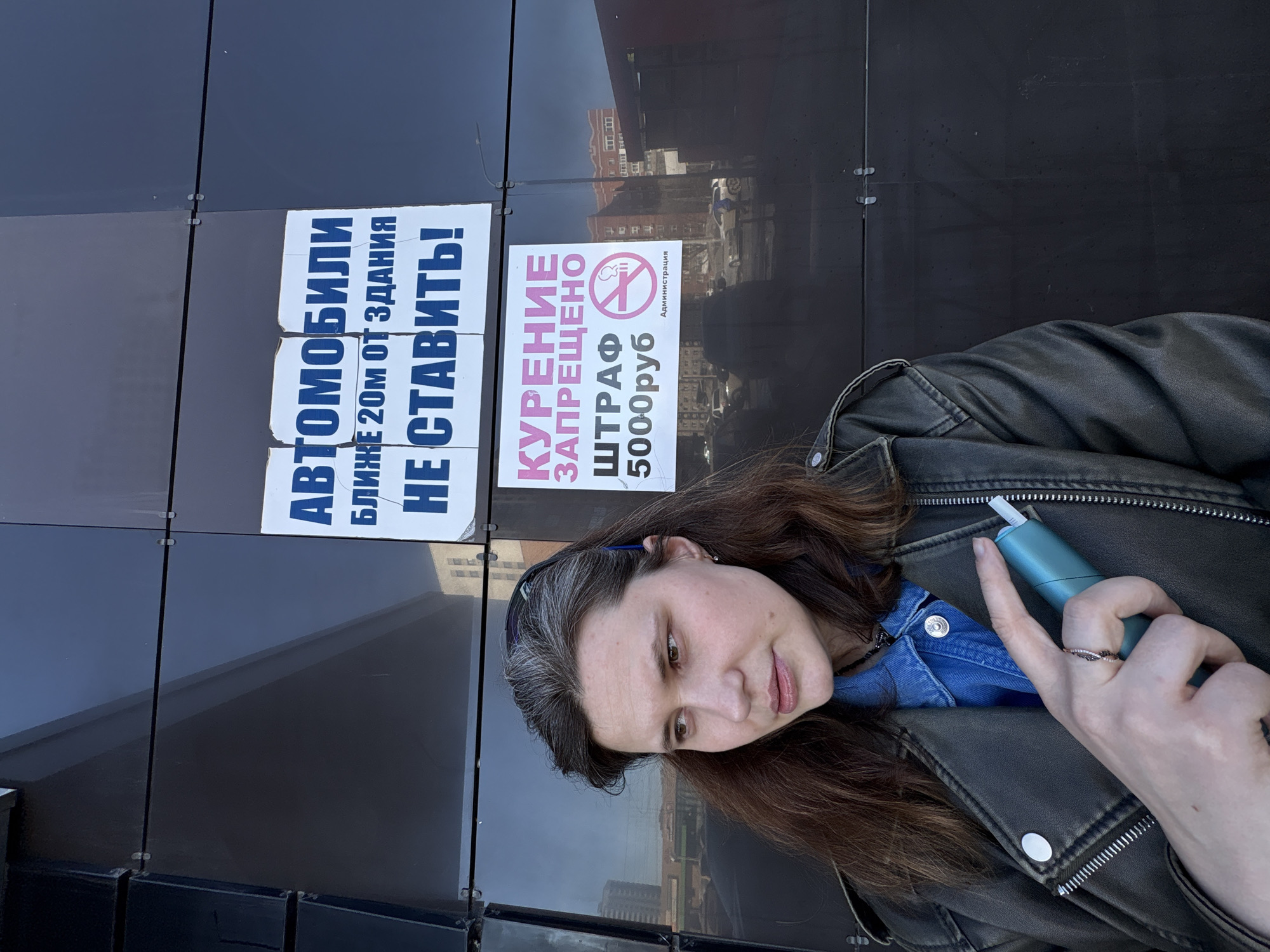 A woman stands in Izhevsk, Russia, positioned in front of a dark, reflective building facade. She has medium-length dark brown hair with visible grey strands and wears a dark, possibly olive-green, leather jacket over a blue denim shirt, along with a thin black choker. A dark headband or sunglasses are pushed up onto her head. Her left hand, adorned with a ring on the ring finger, holds a light teal-colored heated tobacco device with a white stick inserted. Her gaze is directed towards her left.
Affixed to the reflective facade behind her are two white rectangular signs with Russian text. The upper sign, in blue text, reads: "АВТОМОБИЛИ БЛИЖЕ 20М ОТ ЗДАНИЯ НЕ СТАВИТЬ!" (AUTOMOBILES NOT TO BE PARKED CLOSER THAN 20M FROM THE BUILDING!). The lower sign, featuring pink and black text, states: "КУРЕНИЕ ЗАПРЕЩЕНО ШТРАФ 5000руб" (SMOKING IS PROHIBITED FINE 5000 RUB), accompanied by a "no smoking" pictogram and the word "Администрация" (Administration). The reflections in the building facade show a clear sky, a street light, and distant urban structures including a multi-story building and a parking area with vehicles.