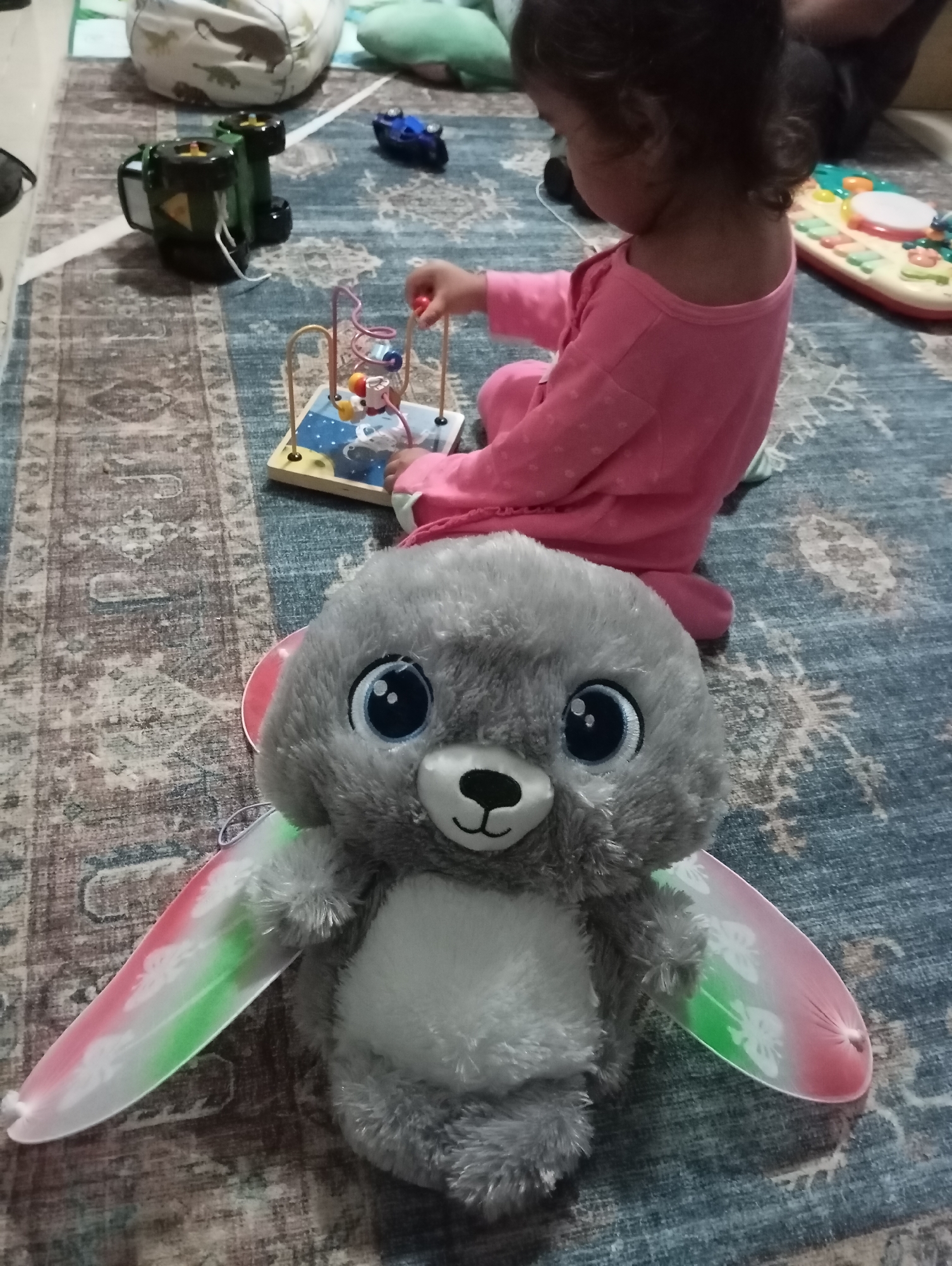 In Dubai, United Arab Emirates, a close-up, eye-level shot captures a soft, gray stuffed animal with large blue eyes, black nose, and a white muzzle. It has translucent, rainbow-colored wings that fade from pink to green, adorned with subtle white butterfly patterns. The stuffed animal is positioned prominently in the foreground, on a patterned blue and gray rug. Behind it, a young child wearing a pink long-sleeved shirt and leggings is partially visible, seated on the rug. The child's hands are engaged with a colorful wooden bead maze toy. Further in the background, a green toy vehicle is visible, along with other blurred children's toys and a large, soft beanbag chair with dinosaur patterns. The overall environment suggests a play area within a home.