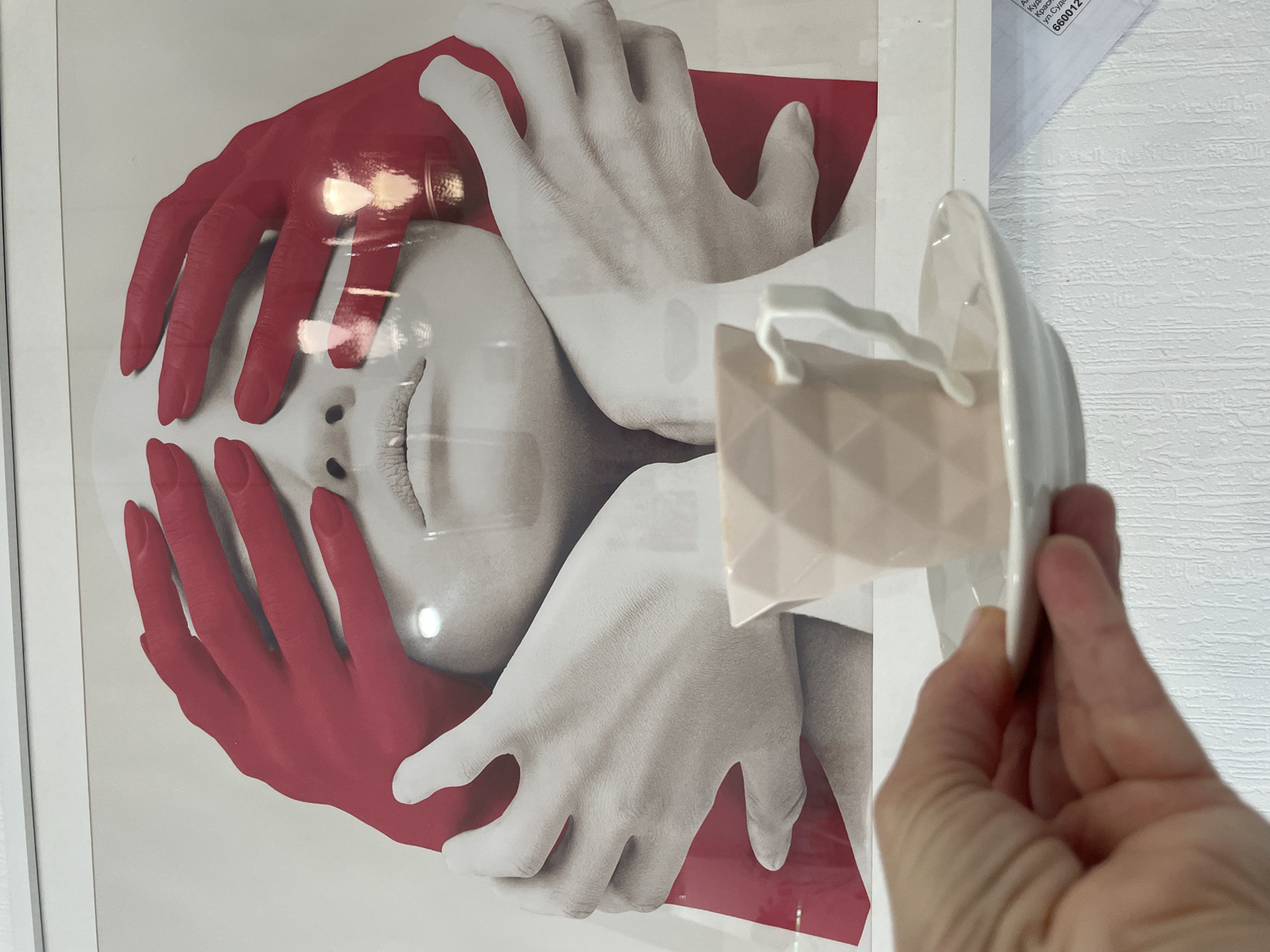The image shows a framed poster of a person's face partially obscured by two pairs of hands, one painted red and the other white. A hand holds a small, geometric-patterned object in front of the poster. An envelope with Russian text including "Krasnoyarsk, ул. Судостр" and a zip code beginning with "660012" is partially visible on the wall above the poster. The location is in the city of Krasnoyarsk, Russia. The wall has a textured, off-white surface.