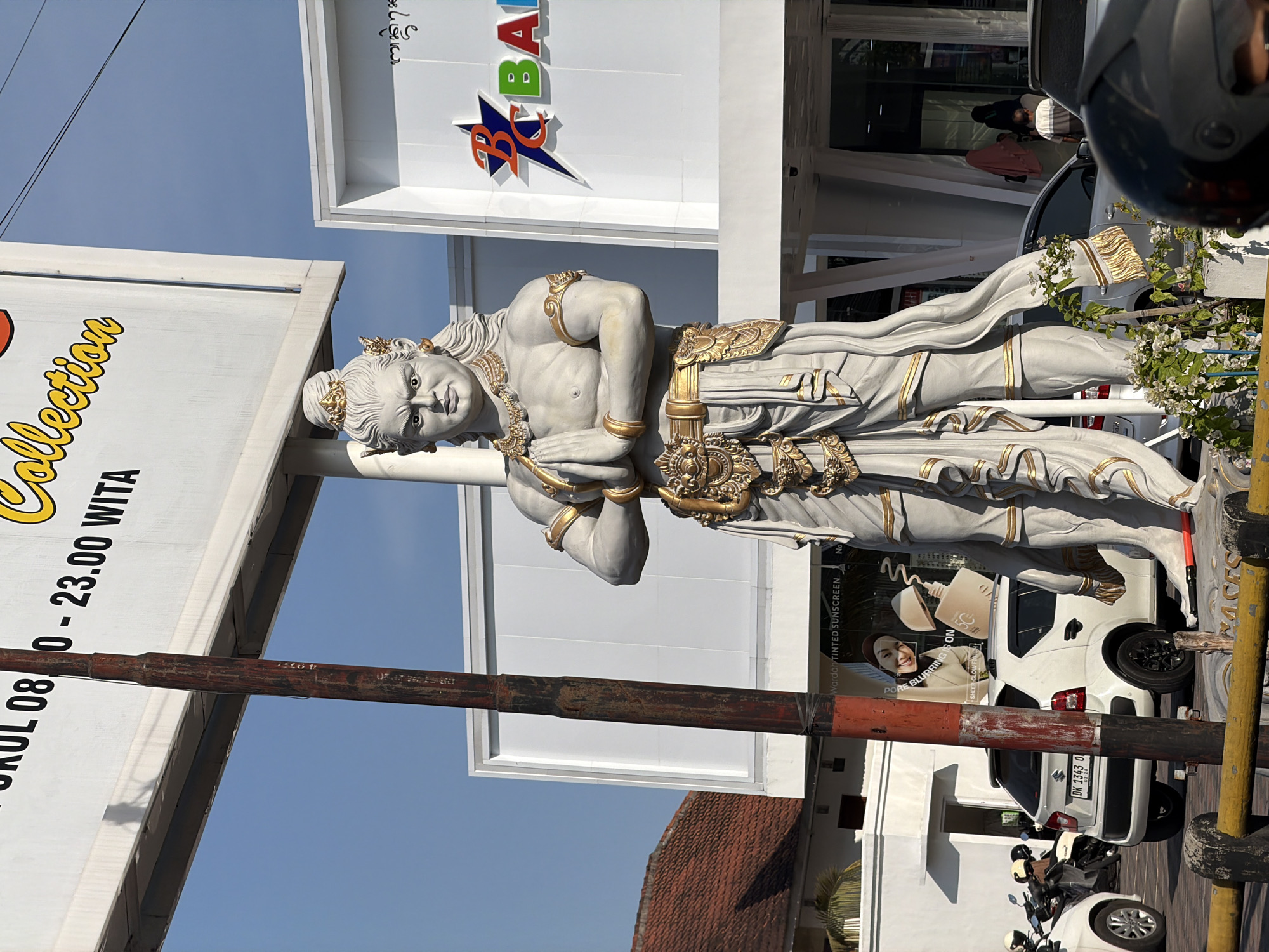 This is a medium shot, eye-level, outdoor, daytime photograph taken in Denpasar, Indonesia. In the foreground, a large, white, stone statue of a male figure with golden accents stands center-frame. The statue is adorned with intricate jewelry, a belt, and draped clothing. Its hands are clasped in front of its chest, and it has a stern expression. To the left of the statue is a rusty, vertical metal pole. Behind the statue and pole, a bright white billboard with black and yellow text is visible. Part of the text reads "Collection" in yellow script and "08:00 - 23:00 WITA" in black. To the right of the statue is a modern building with white walls and large glass windows. A blue and red logo with the letters "BC" and a star is displayed on the building. A white car with a license plate that reads "DK 1343 O" is parked behind a yellow and black barrier in the middle ground. Several motorcycles are parked to the left of the car. The background consists of a clear blue sky and the tiled roof of a building on the left. The environment appears to be a commercial area, possibly a street or plaza.