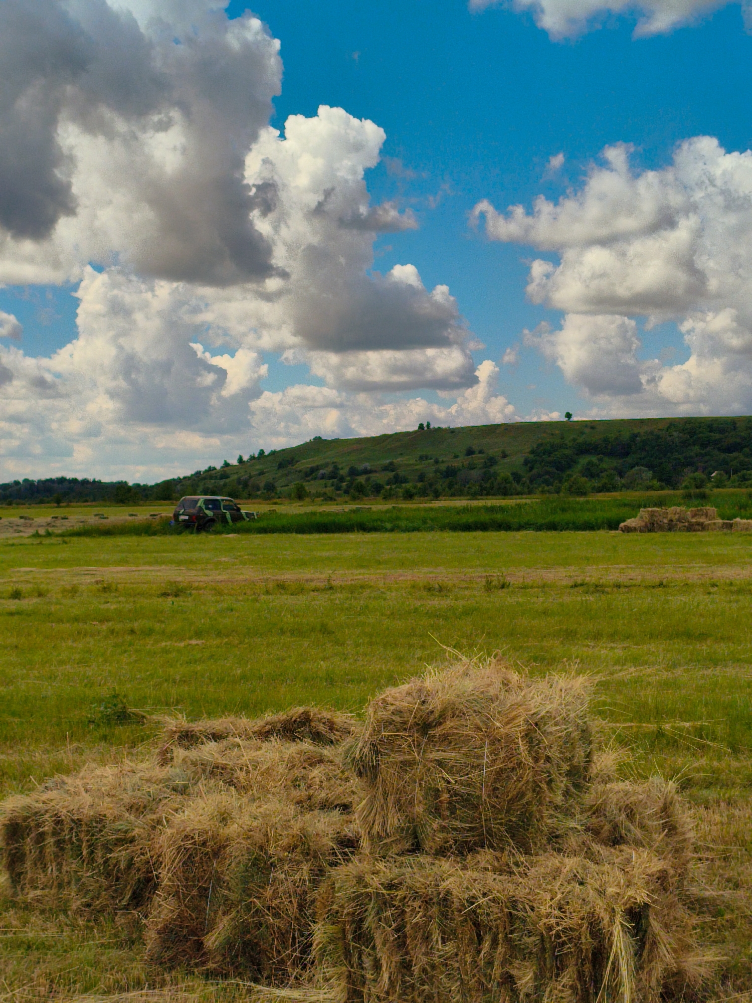 A photograph captures a rural landscape under a bright blue sky dotted with fluffy white clouds. In the foreground, several rectangular hay bales are stacked on a green grassy field. Further back, another collection of hay bales is visible. A dark-colored SUV with a camouflage pattern is parked in a field of green grass. In the distance, a rolling, tree-covered hill forms the backdrop, and a faint treeline runs along the horizon. The scene appears to be located in the village of Ustyvytsia, Ukraine.