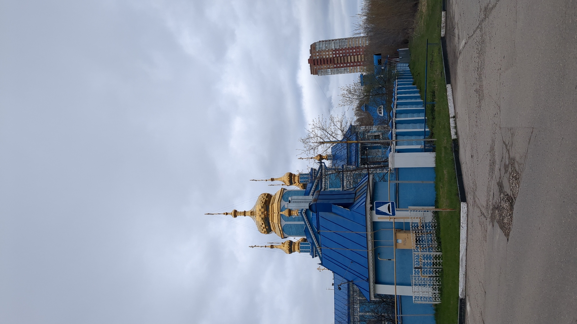 The image displays an urban landscape featuring a vibrant blue Orthodox Christian church with multiple golden onion domes, under an overcast sky. The church, occupying the left and central parts of the frame when oriented correctly (rotated 90 degrees counter-clockwise), boasts intricate detailing on its blue roofs and walls, complementing the shining golden cupolas. Several golden crosses adorn the tops of the domes.
In the background, towards the right side, a tall, multi-story residential building with red and white facades is visible, indicating a developed urban or suburban environment. Various trees, some still bare-branched from winter and others showing early signs of green foliage, are scattered around the church and the background, suggesting a spring season.
In the foreground and to the right, a gray asphalt road with visible cracks runs parallel to a strip of green grass, separated by a white-painted curb. No people or specific events are taking place; the scene is static. The time of day is daylight, characterized by a uniformly cloudy sky, with light grey clouds covering the entire visible expanse.
A notable detail is a blue square sign situated near the church, by the grass strip. This sign features a white upward-pointing triangle, within which there is a smaller black stylized icon resembling a loudspeaker or megaphone. This is likely an informational sign pertaining to the church or its facilities, rather than a standard traffic sign. The location context provided is Ulyanovsk, Russia.