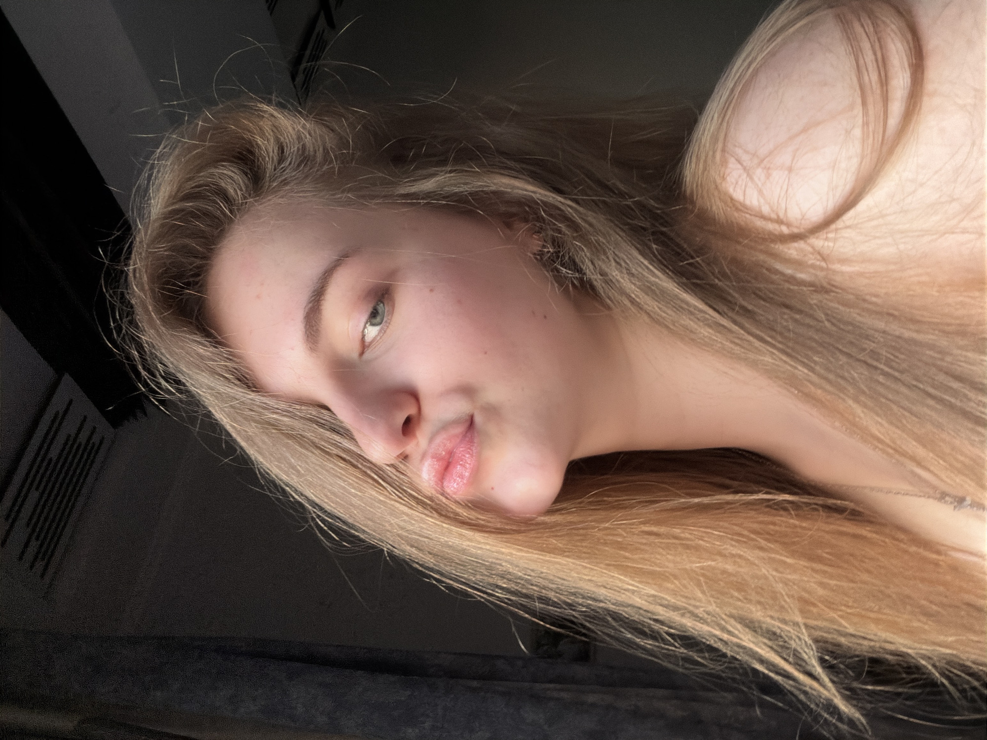 A young woman with long blonde hair is captured in a close-up portrait, looking towards the camera with a playful pout. Her left eye, a striking blue-green, is visible, while her right side is obscured by her hair. The lighting suggests she is near a window, with a dark curtain on the left and a dark ceiling with some sort of textured panels or vents above. The overall mood is casual and intimate. The location is unknown.