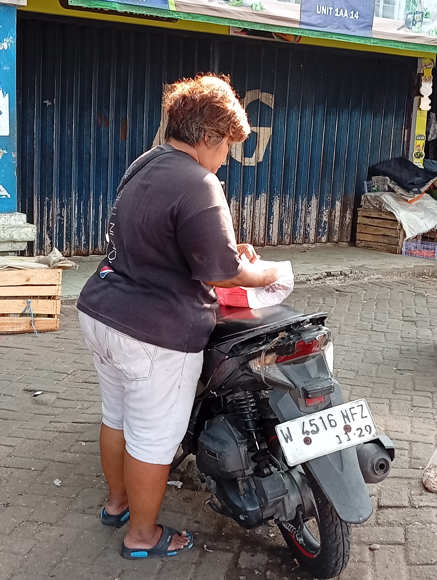 A woman with reddish-brown, permed hair stands next to a parked motorcycle in front of a blue corrugated metal storefront in Larangan, Indonesia. She is wearing a dark gray t-shirt with graphics on the back and light-colored shorts. Her hands are holding a white plastic bag. The motorcycle is dark gray with a license plate that reads "W 4516 NFZ 11.29". Wooden crates are visible to the left of the woman. The ground is paved with brick.