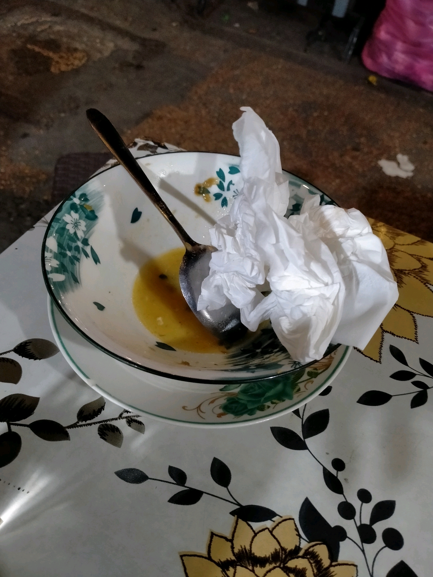 The image depicts a close-up, high-angle shot of a used bowl with a crumpled white tissue on top, resting on a patterned tablecloth. The bowl contains a small amount of yellow liquid. A spoon is partially submerged in the liquid, with its handle extending upwards. The tablecloth has a floral design with large yellow flowers and dark green leaves. The background appears to be a textured, earthy surface, possibly a sidewalk or ground, with a blurred pink object in the upper right corner. The scene suggests a post-meal setting. The location is identified as Ambon, Indonesia, though no specific landmarks or context are provided within the image itself to confirm this.
