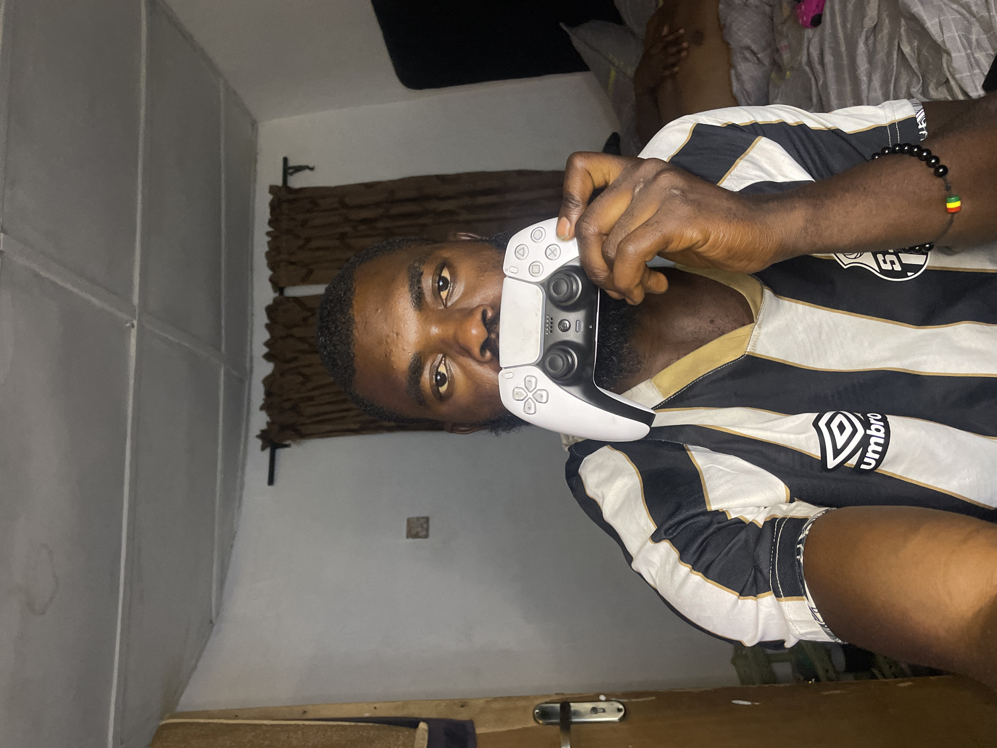A person holds a white PlayStation 5 controller in front of their face, obscuring their mouth and chin. The person has dark skin and a short beard. They are wearing a black and white striped Umbro jersey and a beaded bracelet with a Rastafarian color scheme. The background shows a room with white walls, a dark brown window covering, and a dark couch. The location is likely a residential interior in Jos, Nigeria, given the individual's appearance and style of dress.