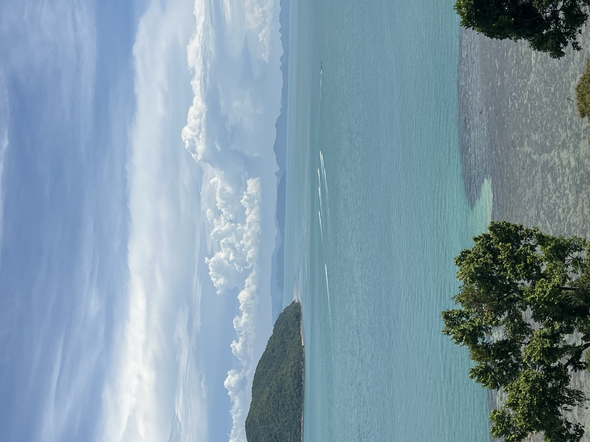 The image depicts a scenic view of the ocean and sky. The sky is a bright blue with streaks of white clouds, suggesting a sunny day. Below the sky, the ocean stretches out, a clear turquoise color. In the distance, a hazy mountain range is visible.
The foreground of the image is dominated by a lush green, tree-covered island, jutting out into the ocean. The water near the island appears shallow and rocky, with visible textures on the seabed. Several small boats are seen moving across the water, leaving white wakes behind them, indicating activity and travel.
On the right side of the image, the branches of a large, leafy tree extend into the frame, partially obscuring the view of the ocean and seabed. The sunlight filtering through the leaves suggests it is daytime.
The overall mood of the image is peaceful and idyllic, evoking a sense of tropical beauty and tranquility. The location cues and the clear, calm water suggest a coastal or island setting, consistent with a tropical environment like Baan Thong Krut, Thailand. No text is visible in the image.