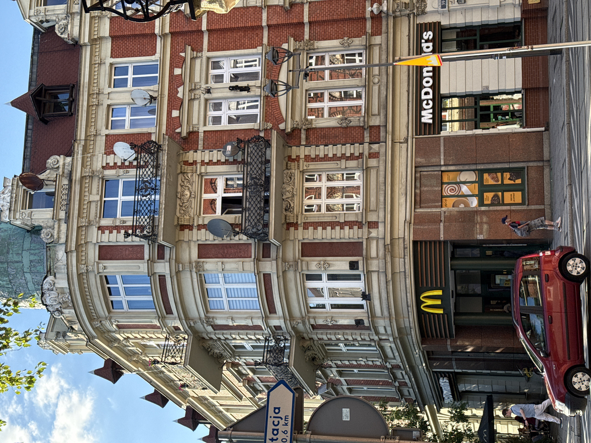 This is a detailed street-level photograph of a multi-story, corner building in a European city, likely from the late 19th or early 20th century, located in the city of Unknown, Unknown. The building features an ornate facade constructed with red brick and lighter-colored (cream or beige) stone or stucco elements, including decorative cornices, window frames, and balustrades.
The building's architectural elements include multiple balconies with intricate black wrought-iron railings. Several white-framed windows are visible across all floors, with some having white blinds. Multiple satellite dishes are affixed to the building's exterior, primarily on the red brick sections. The roof, partially visible, is red-tiled and features several dormer windows, some capped with small, pointed spires. A large, verdigris-patinated dome-like structure crowns the corner section of the building at its highest point.
The ground floor of the building is occupied by a McDonald's restaurant. The distinctive yellow "M" logo is prominent on the corner facade, with "McDonald's" spelled out on the right-hand section of the storefront. A "McCafé" promotional poster is visible in a window on the far right. An outdoor seating area with a black umbrella is partially visible on the far left, in front of another McDonald's entrance.
The scene is set on a sunny day with a clear blue sky showing some scattered white clouds. Sunlight illuminates the building from the left, casting shadows. Green leaves from an overhead tree are visible at the top left of the frame.
On the left side of the image, a blue directional sign attached to a pole reads "Stacja" (Polish for "Station") and "0,6 km," with an arrow pointing to the right. Below it, a round gray sign is partially obscured. A tall black lamppost with two lamps stands in the middle-right foreground, featuring a yellow triangular warning sign attached to it.
Several people are visible on the sidewalk. On the left, a man in a light-colored shirt and trousers walks to the right with his hands in his pockets. Another person is seated at an outdoor McDonald's table, partially obscured by the umbrella. On the right, a man wearing a hat, a dark shirt, camouflage shorts, and a backpack walks towards the left.
A red five-door hatchback car is positioned on the street in front of the building, facing slightly left and away from the viewer. White road markings are visible on the asphalt.