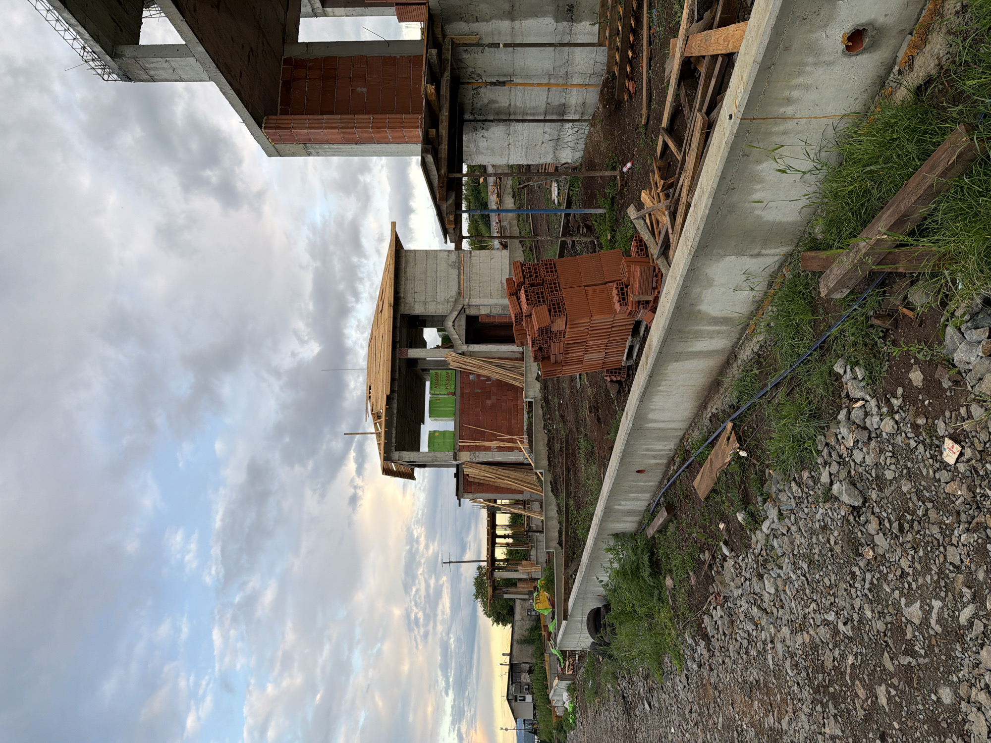 The image shows an ongoing construction site in what appears to be Cherni Vrah, Bulgaria, based on the buildings in the background. Several unfinished concrete and brick structures are visible, suggesting multiple units under construction. Piles of bricks are stacked near the structures. Construction materials, including wooden planks and support beams, are scattered throughout the site. A concrete retaining wall runs along the edge of the construction area, with grass and weeds growing along its base. The ground is covered in gravel and dirt. The sky is overcast with a mix of grey and blue clouds. In the distance, there are houses and power lines. A person wearing a high-visibility vest is briefly visible, further suggesting an active construction site.