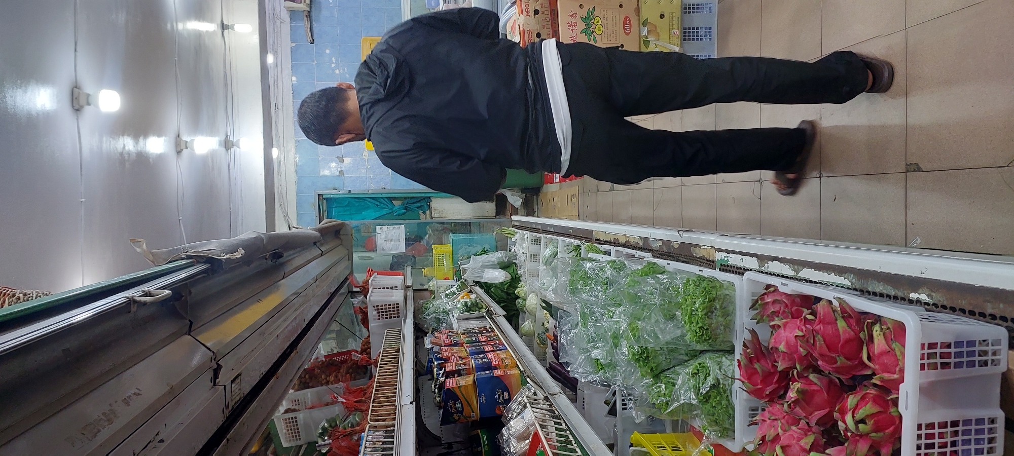 The image depicts a man wearing a black hooded jacket and dark pants, with a white undershirt visible at the waist, walking through a grocery store. He is also wearing brown sandals. The store environment appears to be a small local market, possibly in Jayapura, Indonesia. Shelves are filled with various produce, including dragon fruit in the foreground in white plastic baskets, and pre-packaged vegetables like lettuce in clear plastic bags. There are also boxes of packaged goods on higher shelves and on the floor. The lighting suggests an indoor setting with fluorescent lights on the ceiling. The background shows tiled walls in a light blue color and a partially open teal curtain or door. The man is facing away from the camera and appears to be browsing or shopping.
