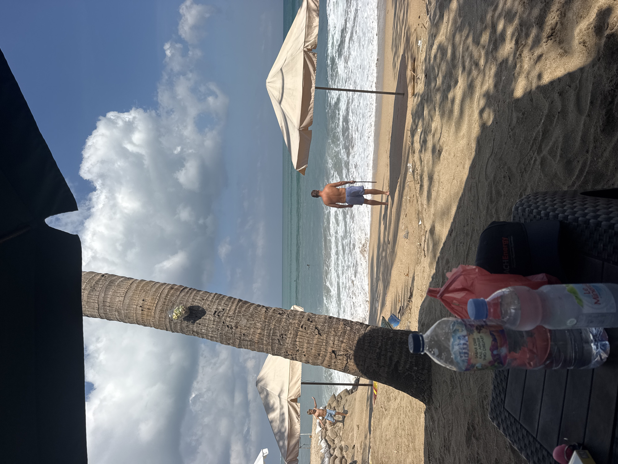 The image depicts a beach scene in Kuta, Indonesia. In the foreground, a table holds two plastic water bottles and a red plastic bag. A black baseball cap with the "ACH Energy" logo is also on the table. The table is positioned on a sandy beach, partially shaded by a large palm tree on the left.
Further into the scene, the ocean is visible with waves crashing onto the shore. Two beach umbrellas are set up on the sand. One umbrella is to the left, with a young boy standing near it. The other umbrella is to the right, and an adult male, wearing blue shorts, stands in front of it, facing the ocean and holding a stick. The sky is blue with scattered white clouds. The environment suggests a sunny day at the beach.