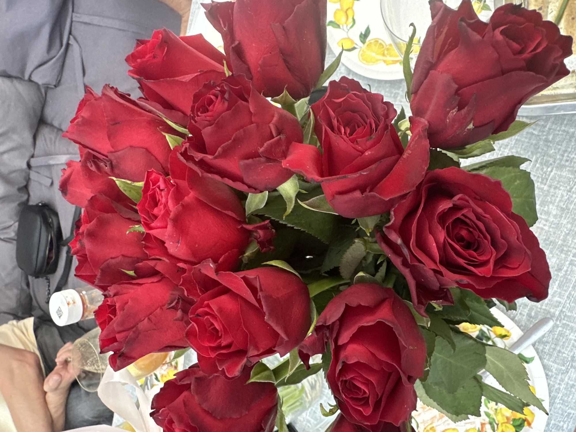 An overhead shot of a bouquet of deep red roses. The roses are densely packed, with some in full bloom and others still in bud. Green leaves are visible amongst the petals. In the background, out of focus, are two hands, one holding a glass of champagne, and a small plastic container with a white lid. A patterned plate with lemons is also visible. The scene appears to be indoors, possibly in a home setting, as indicated by the furniture and general ambiance. No specific landmarks or details are present that would suggest a particular location in Minsk, Belarus.
