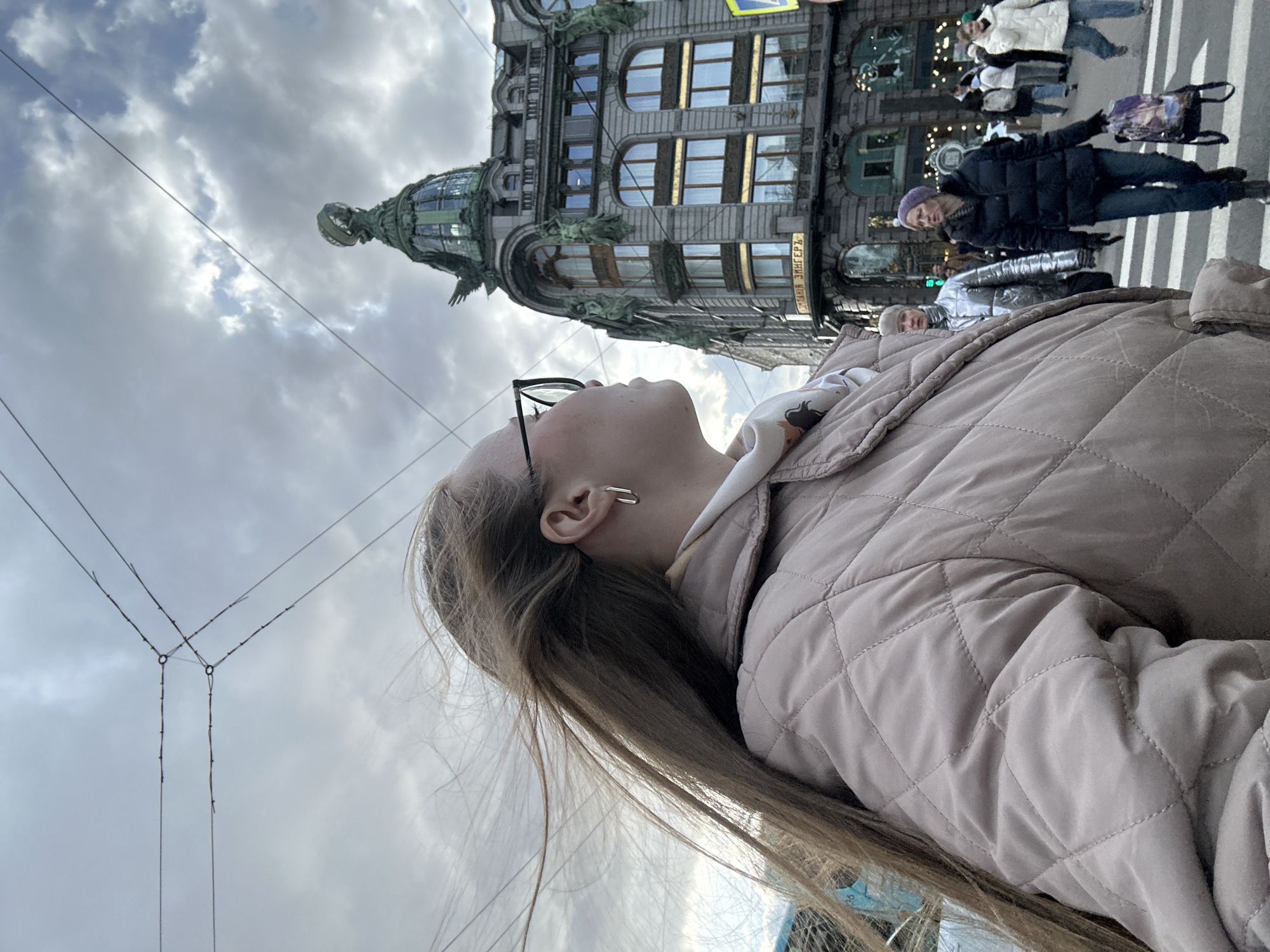 A low-angle, portrait-oriented photograph captures a street scene in Saint Petersburg, Russia, focusing on a young person looking upwards at a grand historic building. In the foreground, a person with long, light brown hair is seen in profile from the chest up, wearing glasses and a light beige quilted jacket. A silver hoop earring is visible in their left ear. They are looking up towards the overcast sky and the ornate architecture.
Dominating the mid-ground is the iconic Singer House (Dom Zingera), a multi-story Art Nouveau building. Its elaborate facade features large windows, classical details, and green decorative elements. A distinctive domed corner tower, topped with a large, statue-like figure, rises towards the sky. Visible on the building's facade is the Cyrillic text "ДОМ ЗИНГЕРА", identifying it as the Singer House.
In the background, several pedestrians are visible on a street or sidewalk in front of the building. One person in a dark coat and purple hat walks across a zebra crossing, carrying a bag. Other figures are further in the distance. Overhead, a network of dark wires, likely for trams or trolleybuses, crisscrosses against the light grey, cloud-filled sky. The scene depicts a bustling urban environment on a cloudy day.