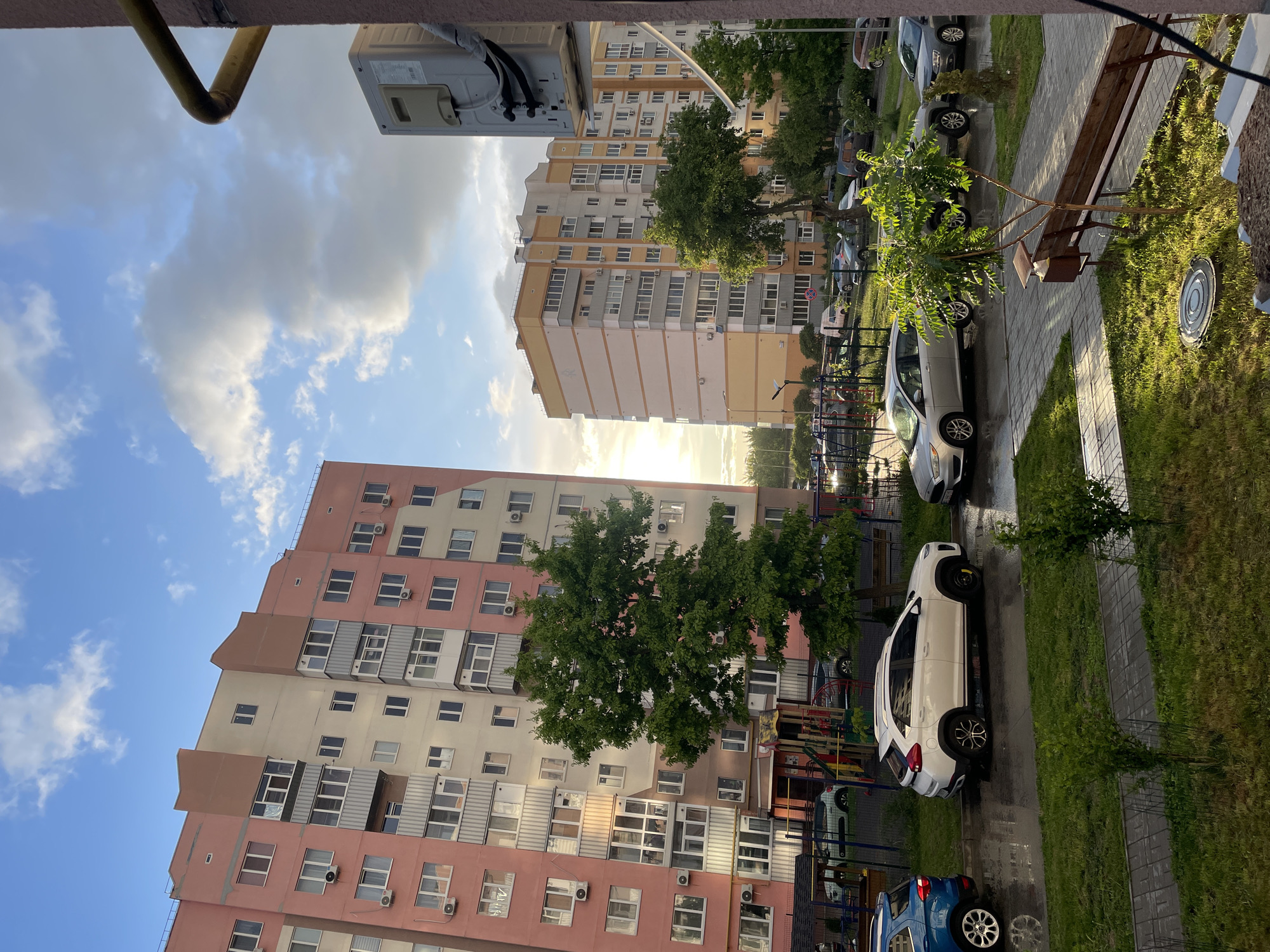 The image depicts an exterior view of a residential area in Rozsoshentsi, Ukraine. Two multi-story apartment buildings are visible, one primarily pink with off-white accents and the other primarily yellow. The buildings appear to be modern, with multiple windows and balconies. Sunlight streams between the buildings, suggesting either sunrise or sunset.
A paved walkway runs through the courtyard between the buildings, with grass and trees lining the sides. Cars are parked along the walkway, including a white SUV and a blue compact car. A wooden bench sits along the edge of the walkway. The overall impression is of a typical urban residential scene.