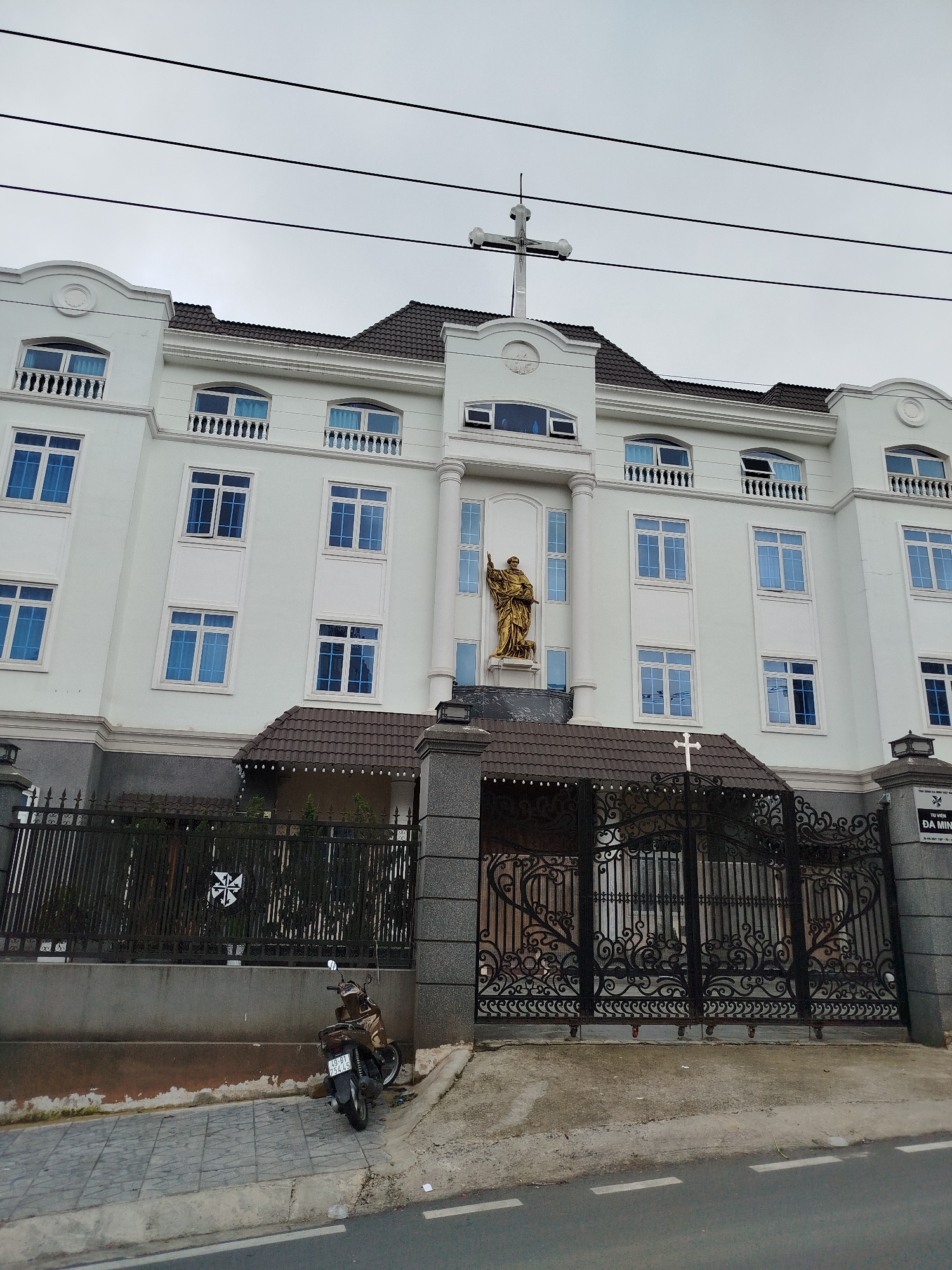 The image displays the facade of a large, multi-story white building, identified as Tu Vien Da Minh (Dominican Monastery/Convent), under an overcast sky during the daytime in Da Lat, Vietnam.
The building features a classical architectural style with multiple rectangular windows, many appearing to have blue panes or curtains. Several windows on the upper floors are accompanied by small balconies with white balustrades. A prominent golden statue of a saint stands in a central, recessed alcove on the building's facade, just above the main entrance. The building is topped with a large white cross, and its roof is dark brown.
In front of the building, an ornate black wrought-iron fence and gate with intricate scrollwork separate it from the street. A smaller white cross is fixed atop a section of the gate. To the right of the gate, a stone pillar bears a sign with the text "TU VIEN" and "DA MINH". Below this, smaller, unreadable text is visible.
A brown and black scooter is parked on the paved sidewalk to the left, displaying a license plate number "49-B1 752.45". Overhead, several power lines stretch across the grey sky. No people are visible in the scene, and no specific activity is taking place, presenting a static view of the building and its immediate surroundings.