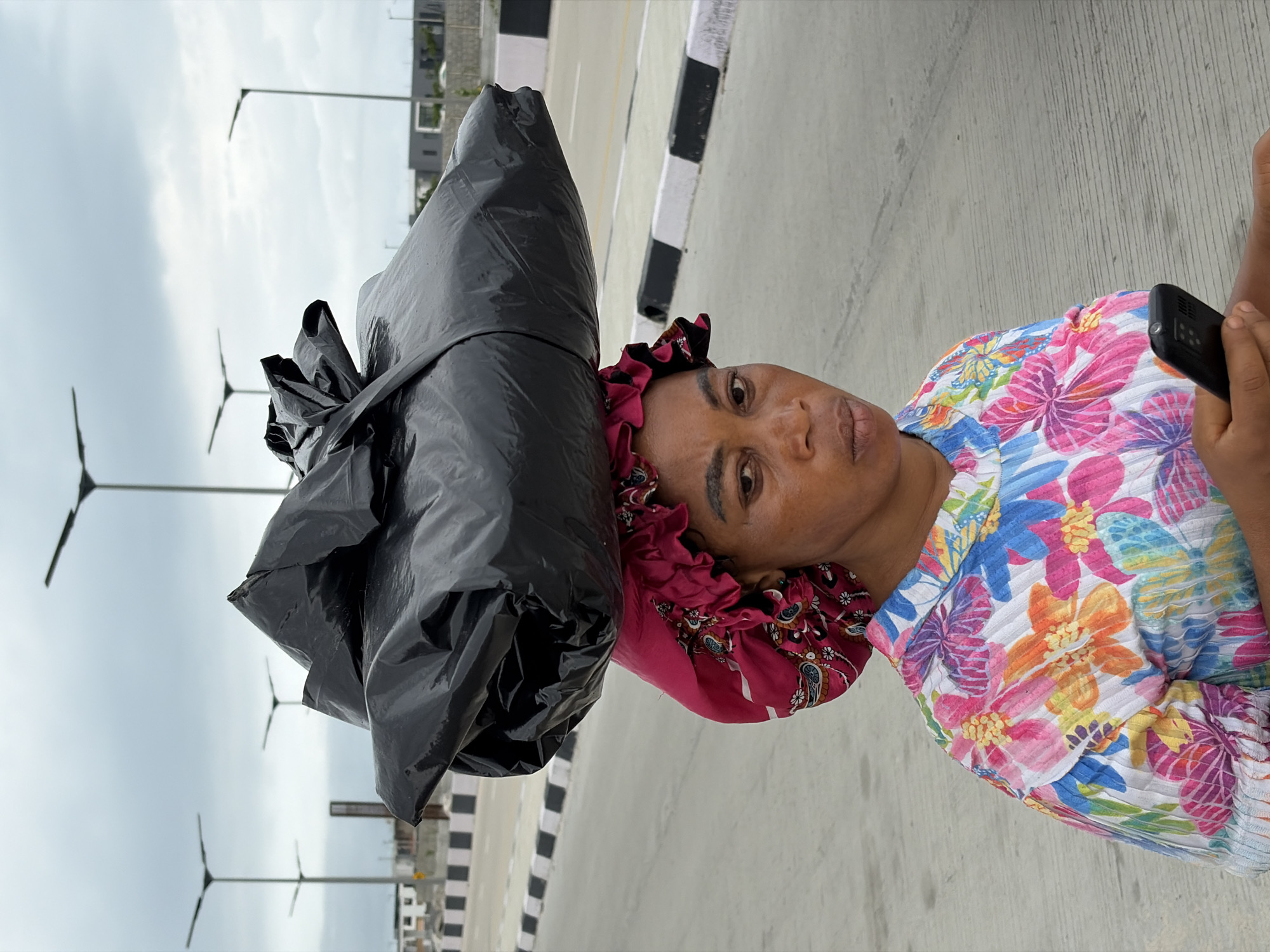 A woman with dark skin, wearing a colorful floral print shirt and a pink headscarf, balances a large black plastic bag on her head. She is walking on a paved road, which appears to be part of a highway or major street in Igboefon, Nigeria. The road is bordered by a concrete barrier with black and white markings. In the background, several modern streetlights with multiple lamps are visible, along with some buildings under construction and other infrastructure elements suggesting a developing urban area. The sky is overcast. The woman is holding a black smartphone in her right hand.