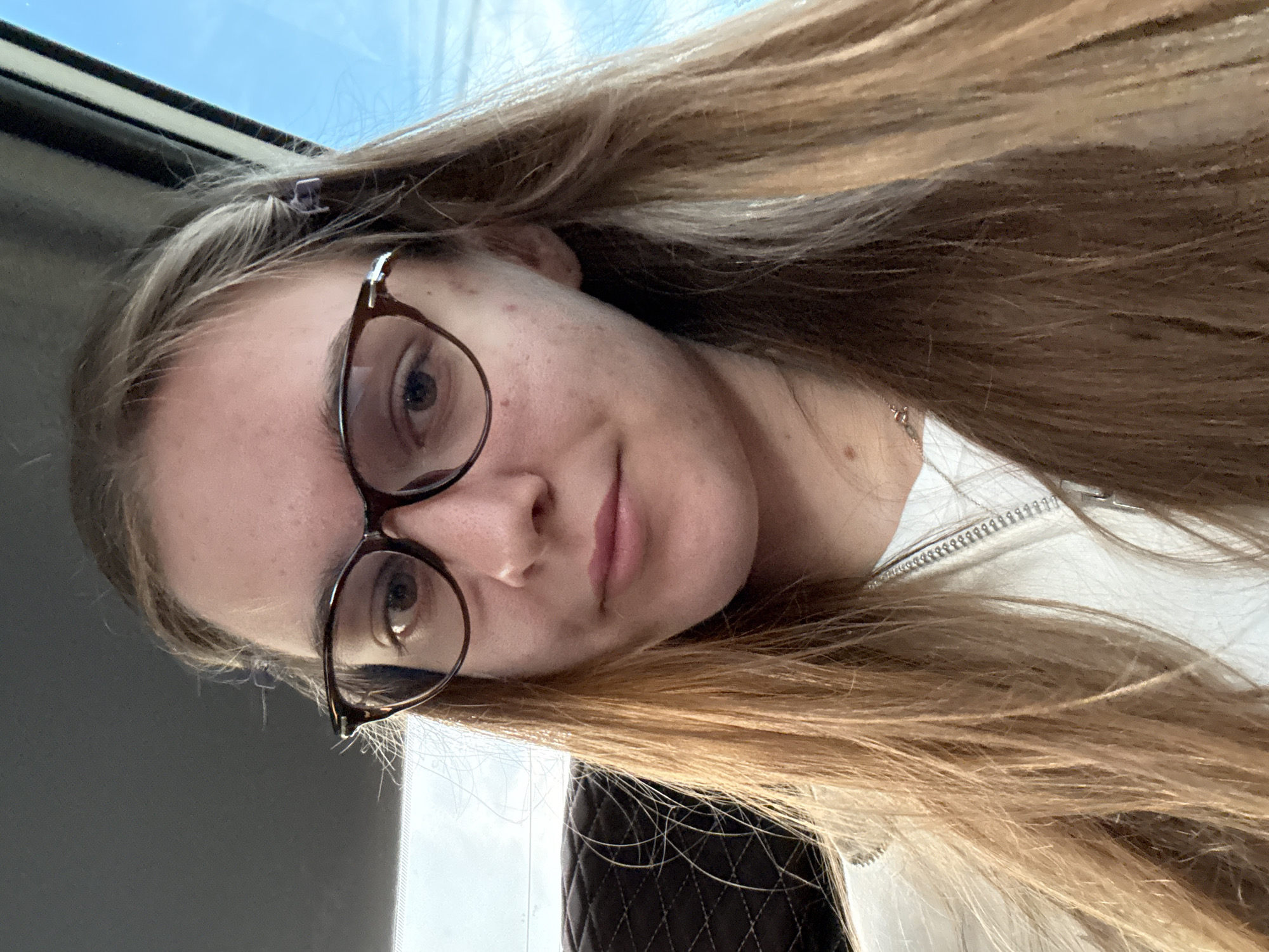 An outdoor, waist-up, eye-level shot shows a fair-skinned young woman with shoulder-length blonde hair, wearing glasses. The woman is looking into the camera, and her face is slightly angled to the right. She has on a white jacket with a zipper. The background is blurred, but it appears to be a car interior, with a dark seat to the left and a window showing a bright blue sky to the right. The woman is in Chaikovsky, Russia.