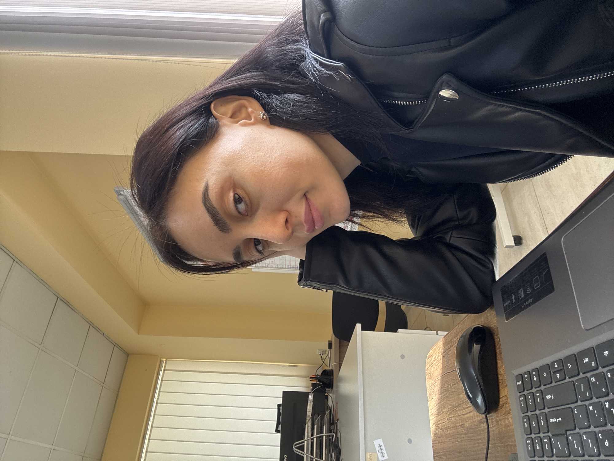 A woman is sitting at a desk, wearing a black leather jacket. She has dark hair and eyebrows. On the desk are a laptop and a mouse. The room appears to be an office with a white ceiling and blinds on the window. A white cabinet and a black chair are visible in the background. It is not possible to determine the location in Tbilisi, Georgia from this image.