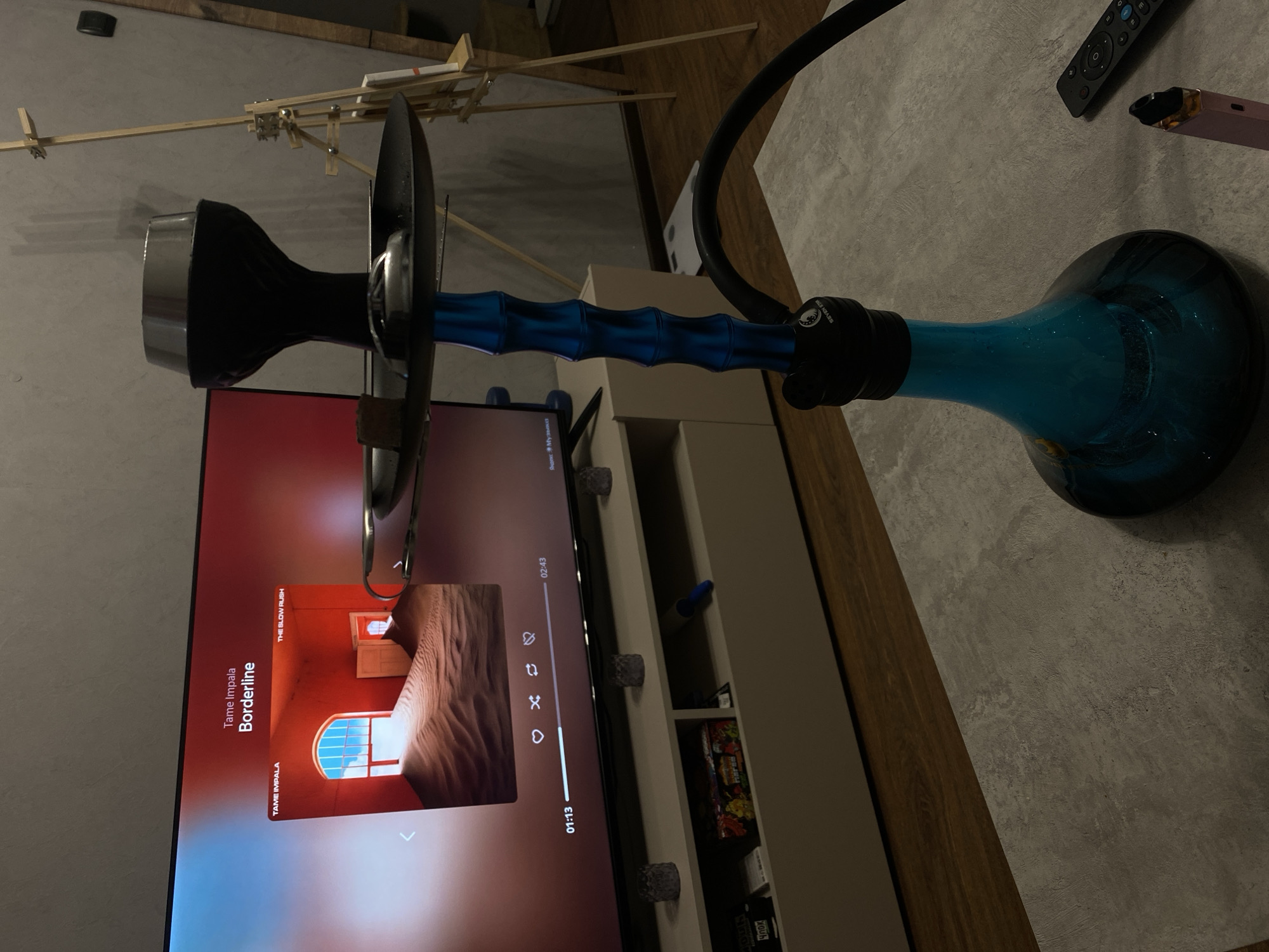 The image shows a living room setting. Dominating the foreground is a blue hookah, positioned near a television displaying the song "Borderline" by Tame Impala. The TV sits atop a beige shelf, and the background includes a wooden structure, possibly a decorative piece or a partially constructed object. A remote control and a vape device are situated on a gray surface in the upper right corner. The scene doesn't offer clear details that would indicate a specific location within Izhevsk, Russia.