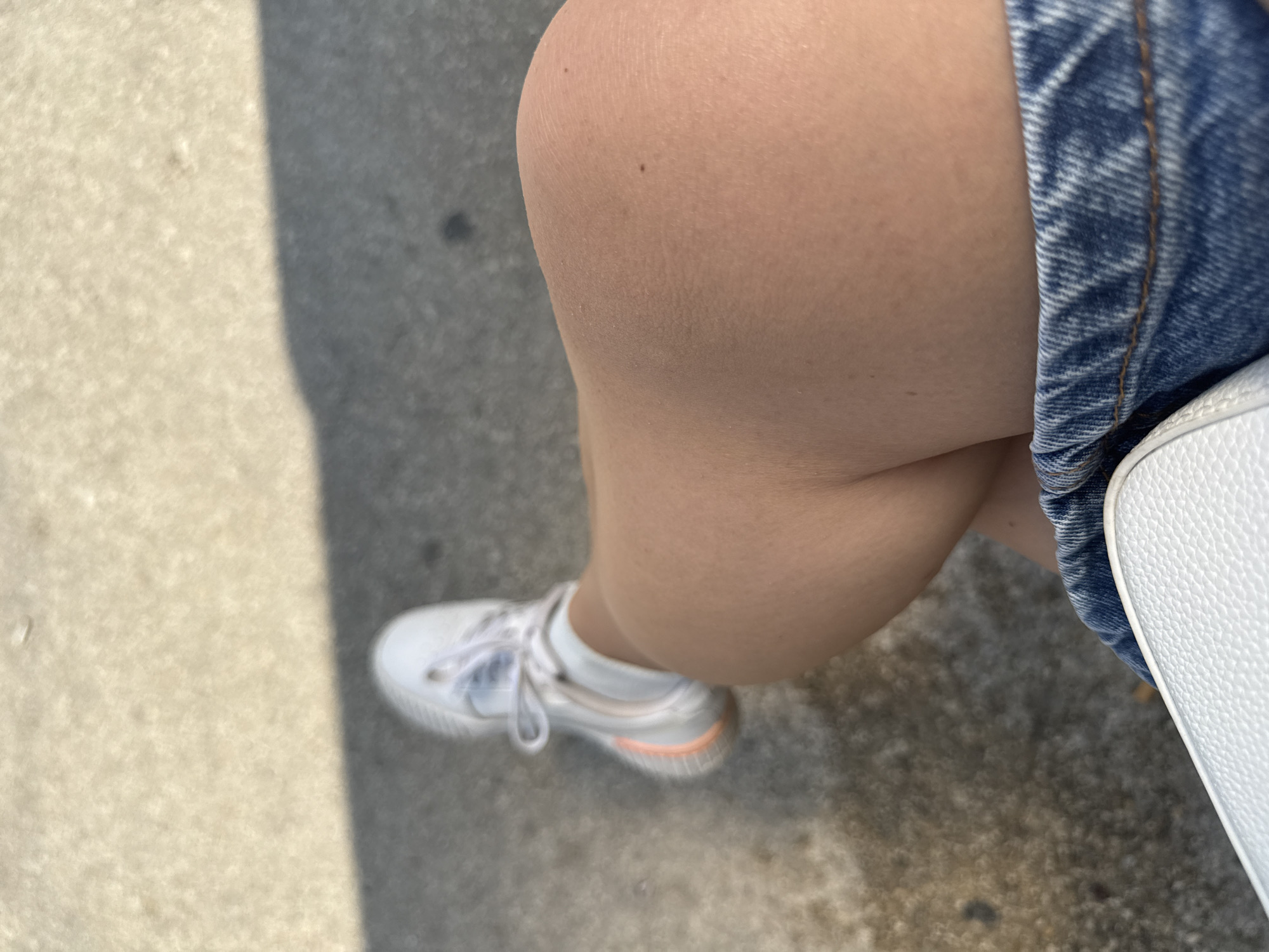 The image, taken from a high-angle perspective, captures a seated individual's leg, foot, and immediate surroundings. A tanned human leg, bent at the knee, occupies the center-right of the frame, exhibiting minor skin texture and small moles. The left foot is shod in a white athletic shoe with white laces, a pale blue detail near the laces, and a distinct peach-colored outsole visible on the medial side. The individual is wearing blue denim fabric, consistent with shorts or a skirt, visible along the bottom and lower-right edges, featuring a coarse weave and orange-brown stitching. A white, textured, possibly synthetic leather bag is partially visible along the bottom edge. The ground beneath is composed of concrete, bisected diagonally by a sharp shadow line from the upper-left to the lower-right. The concrete in sunlight is light-colored and appears dry, while the concrete within the shadow is darker and exhibits some visible moisture or stains. The overall lighting suggests strong, direct sunlight.