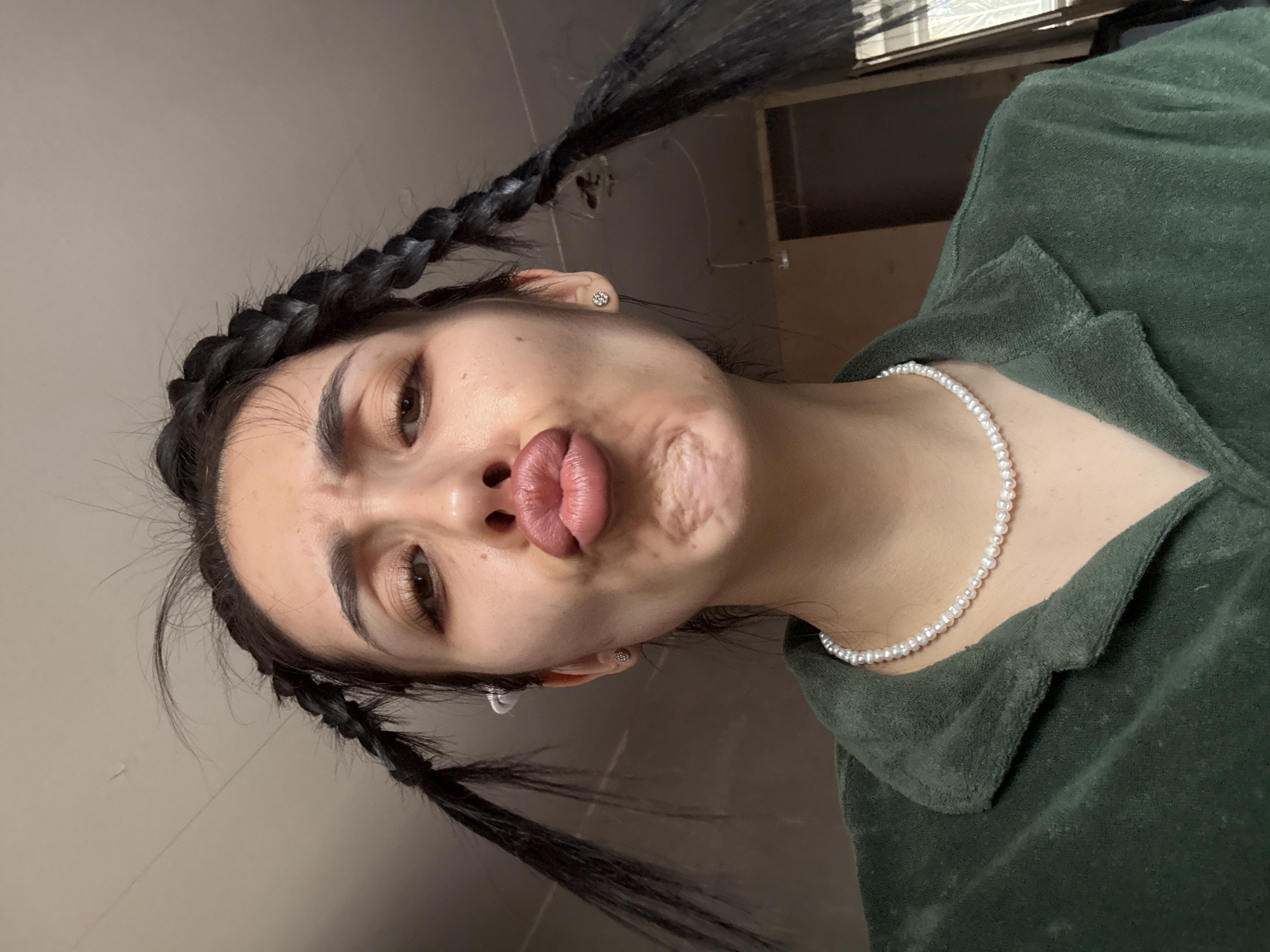A close-up portrait shows a young person, likely female, in an indoor setting in Votkinsk, Russia. The image is rotated, presenting the subject's head tilted with the camera looking slightly up at them. The person has long, dark hair styled into two prominent braided pigtails. They are making a playful "duck face" or "fish face" expression, with lips pursed outwards towards the camera.
The subject wears a delicate pearl choker-style necklace and small, silver-colored stud earrings. They are dressed in a dark green, textured garment, possibly a fleece or velour top or jacket, featuring a visible collar. Notable details include their dark eyebrows and brown eyes. Some natural skin texture and minor imperfections are visible on the face, particularly on the cheeks.
The background is indistinct, appearing as a plain, light-colored wall behind the head and a darker, ambiguous area with faint structural lines (possibly furniture or a door frame) behind the shoulder. The lighting is soft and even, suggesting an indoor environment. There is no visible text. The overall impression is one of casualness and humor, stemming from the subject's expressive pose.