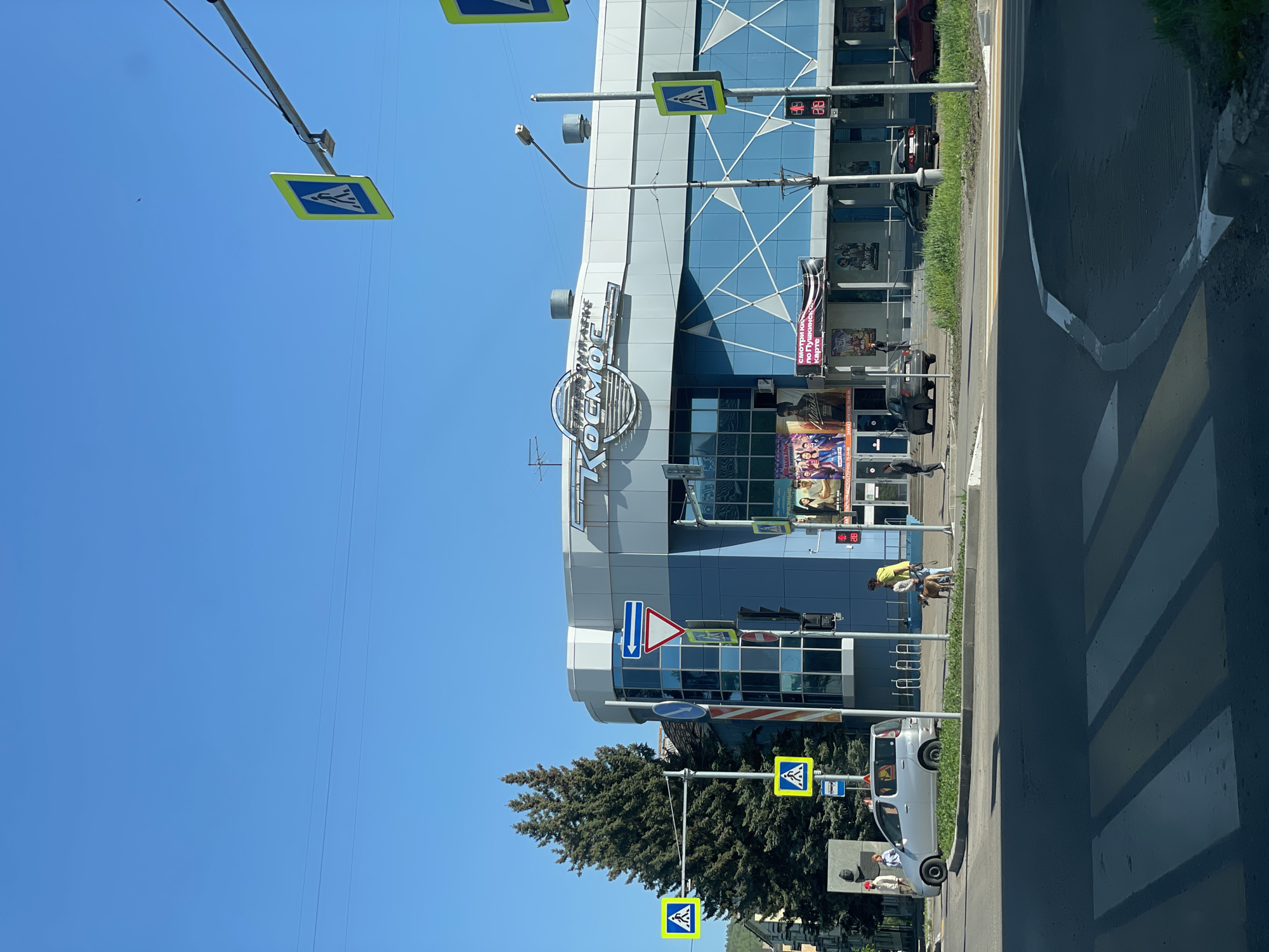 The image captures a vibrant street scene in Zheleznogorsk, Russia, on a bright, clear day with a prominent blue sky. The focal point is a modern cinema building named "КОСМОС" (Kosmos), featuring a striking blue and grey facade with geometric window patterns. Below the main "КИНОТЕАТР" (Kinoteatr - Cinema) sign, various movie posters are displayed, including one for "ДИПЛОМ 2" and a banner promoting "СМОТРИ КИНО ПО ПУШКИНСКОЙ КАРТЕ" (Watch movies with Pushkin Card), indicating a cultural payment method.
In the foreground, a clearly marked pedestrian crossing with white zebra stripes spans the asphalt road. Numerous yellow square signs with black pedestrian symbols signify the crossing, and traffic lights are visible, with one displaying a "08" countdown, likely for pedestrians or vehicles. Other road signs include a blue rectangular sign with a white arrow pointing downwards and a round blue sign with a white arrow pointing right, indicating traffic directions.
Several people are visible in the scene, including an individual in a yellow top near the building's entrance, and others walking near the street. Two cars, a silver hatchback and a darker sedan, are parked along the curb. To the left of the building, a large evergreen tree stands tall, and a monument with a bust is partially visible. Thin overhead wires stretch across the clear blue sky. Bicycle racks are also present near the building entrance.